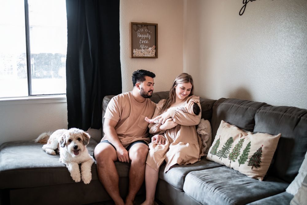 Couple relaxes on grey couch with dog while holding infant in cozy living room setting.