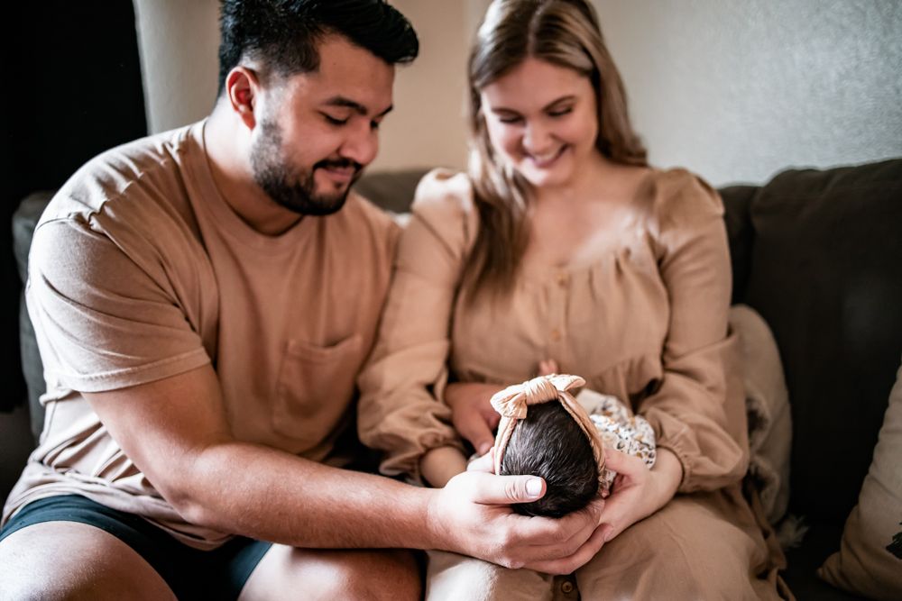 A couple in matching beige outfits sits on a sofa holding a small baby.
