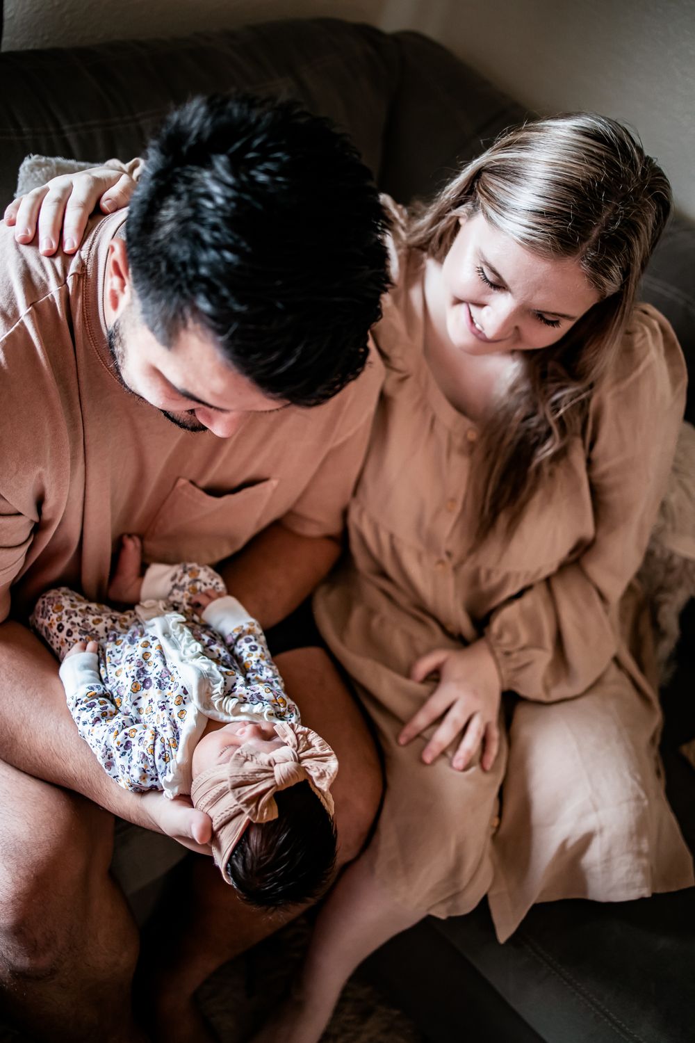 A couple in matching beige outfits shares an intimate moment with their newborn on a gray couch.
