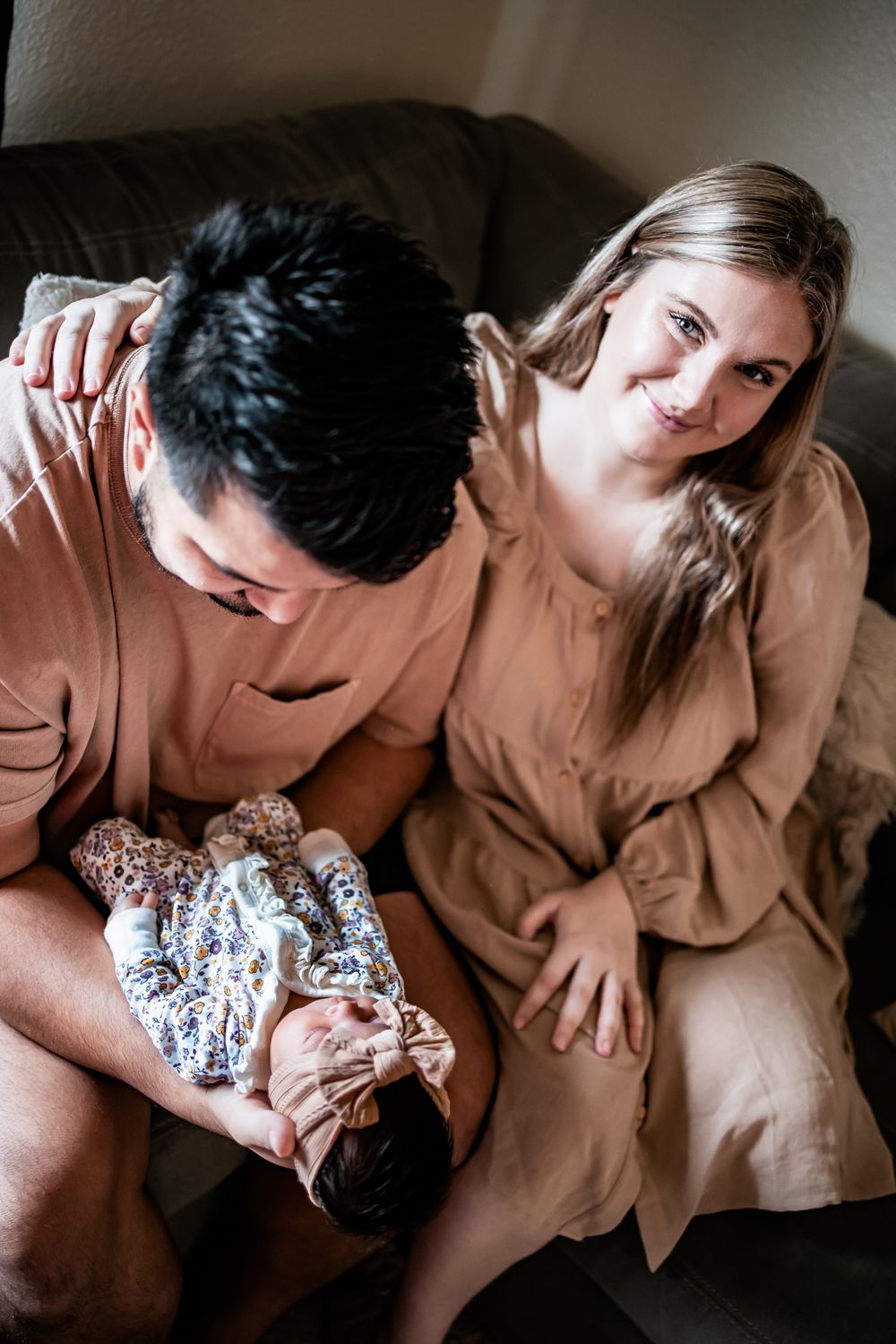 A couple in beige clothing cuddles with their newborn on a comfortable gray couch.