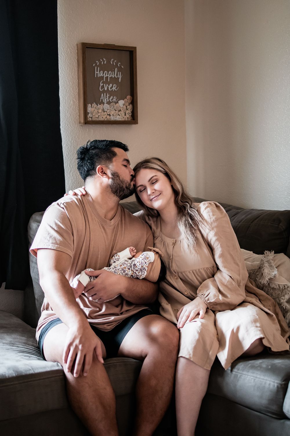 A couple in coordinating beige outfits shares a tender moment on their sofa beneath a wall sign.