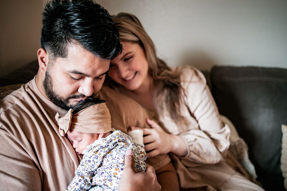 A couple in neutral tones shares a tender moment with their baby on a gray sofa.