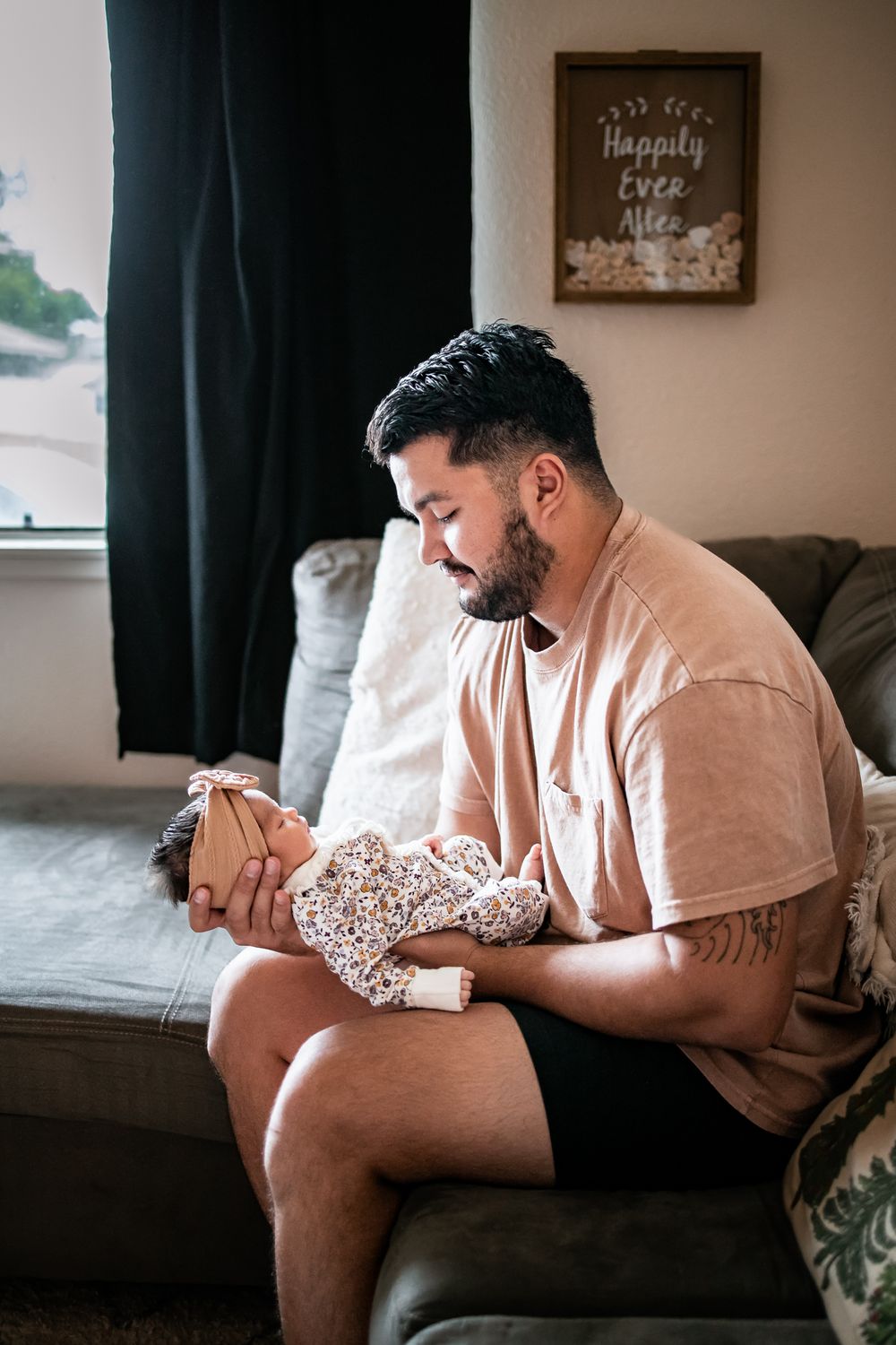 Parent sits on couch holding sleeping infant dressed in floral print onesie.