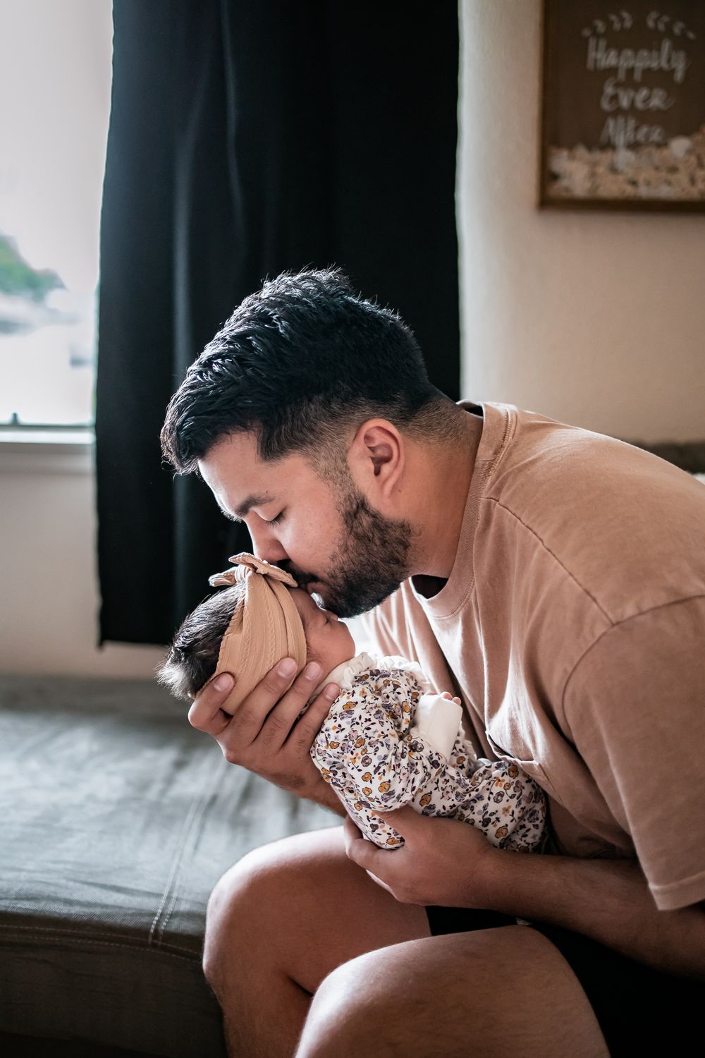 Parent in peach shirt tenderly cradling and kissing newborn baby in floral outfit on gray couch.