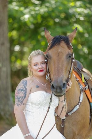 A Bride in a white wedding dress poses with a brown horse wearing in a sunlit wooded setting.