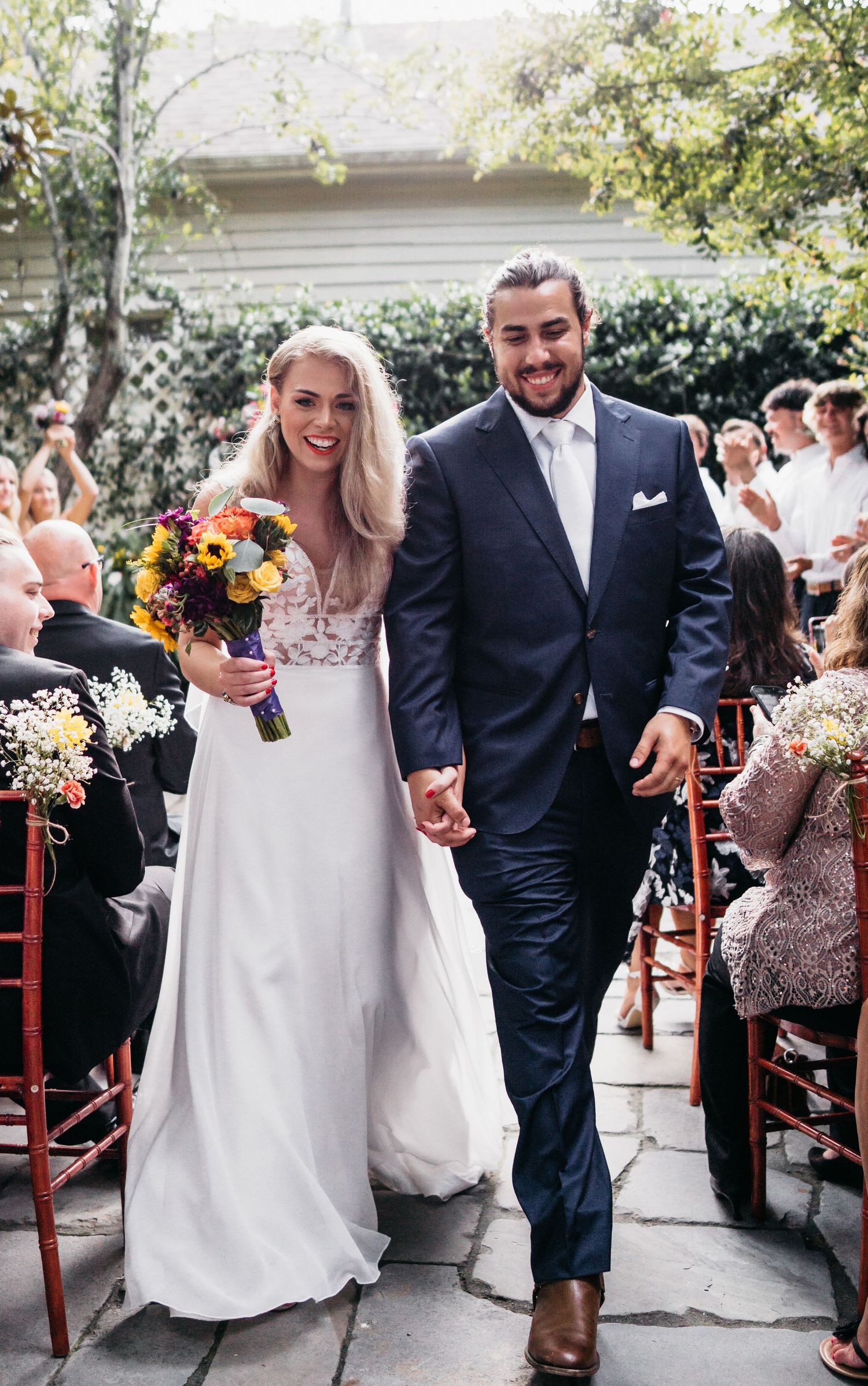 A bride in a white wedding dress holding a colorful bouquet and her grooms hand walk down the aisle together smiling