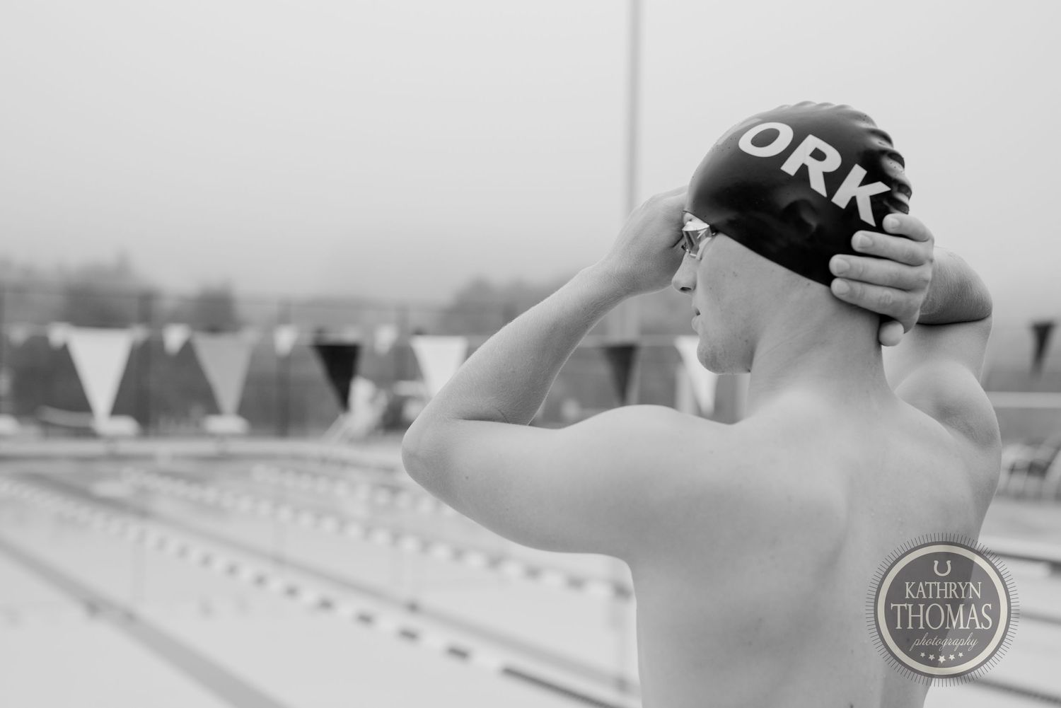 A swimmer adjusts their cap while standing at the edge of a pool in black and white.
