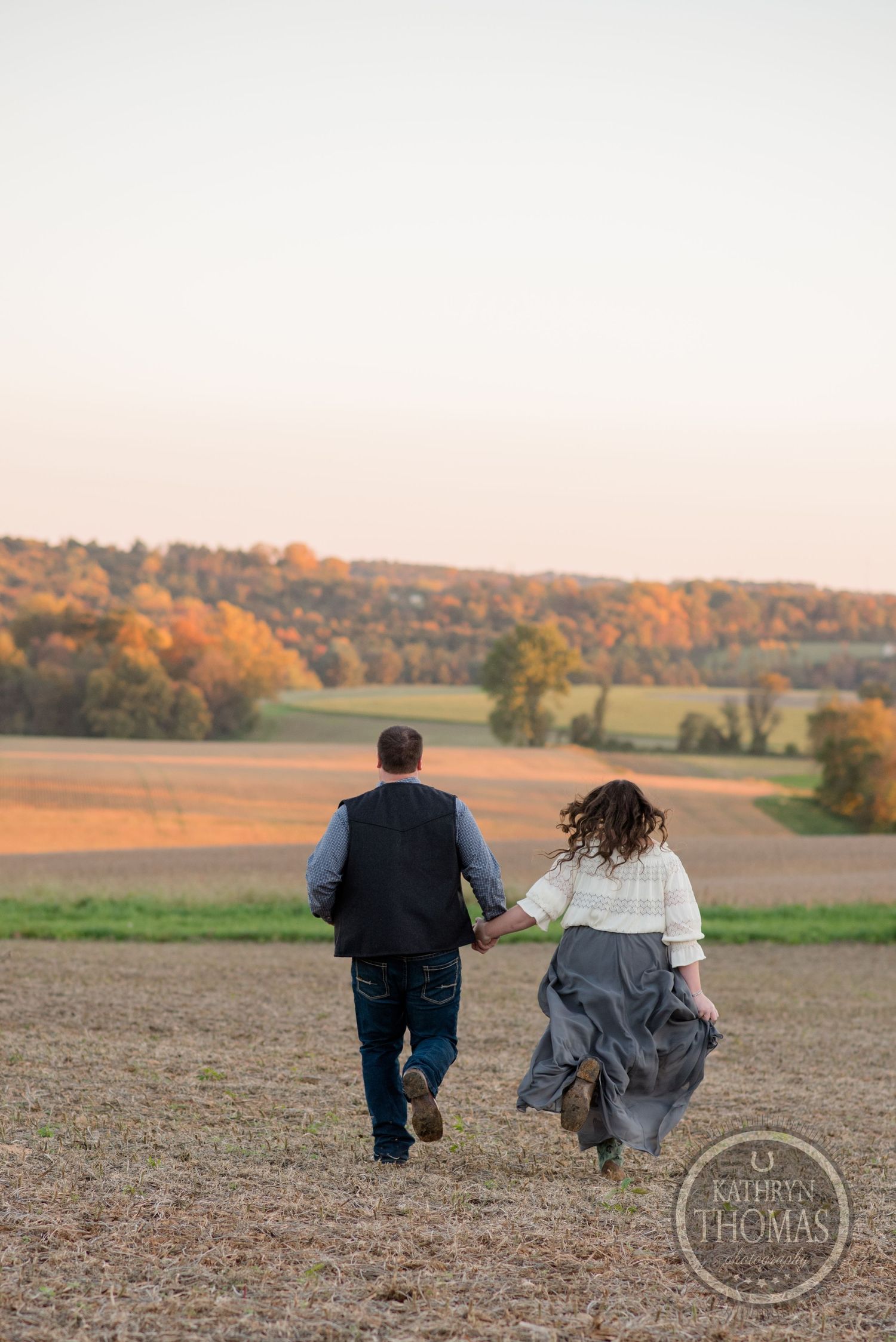 Couple walking hand in hand through an open field at sunset with rolling hills in the distance.