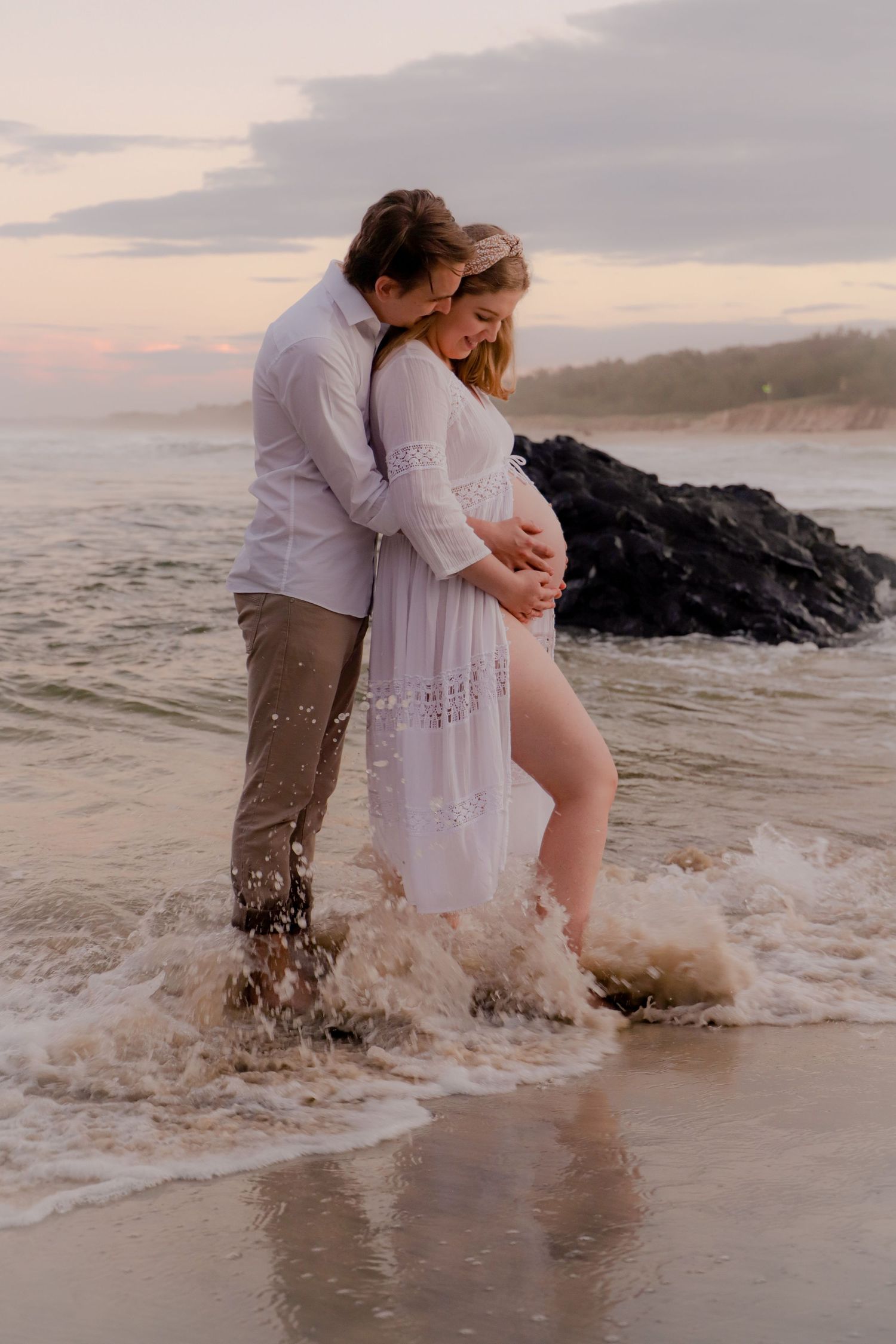 Romantic couple embracing in ocean waves during sunset wearing white and beige outfits.