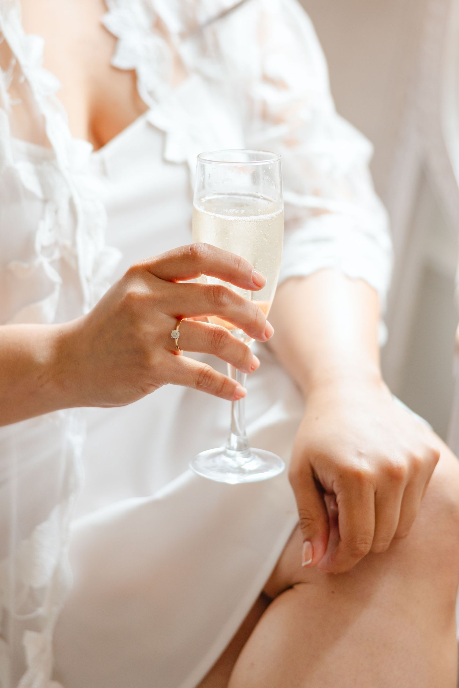 Close-up of bride in white robe holding champagne flute with engagement ring visible on hand.