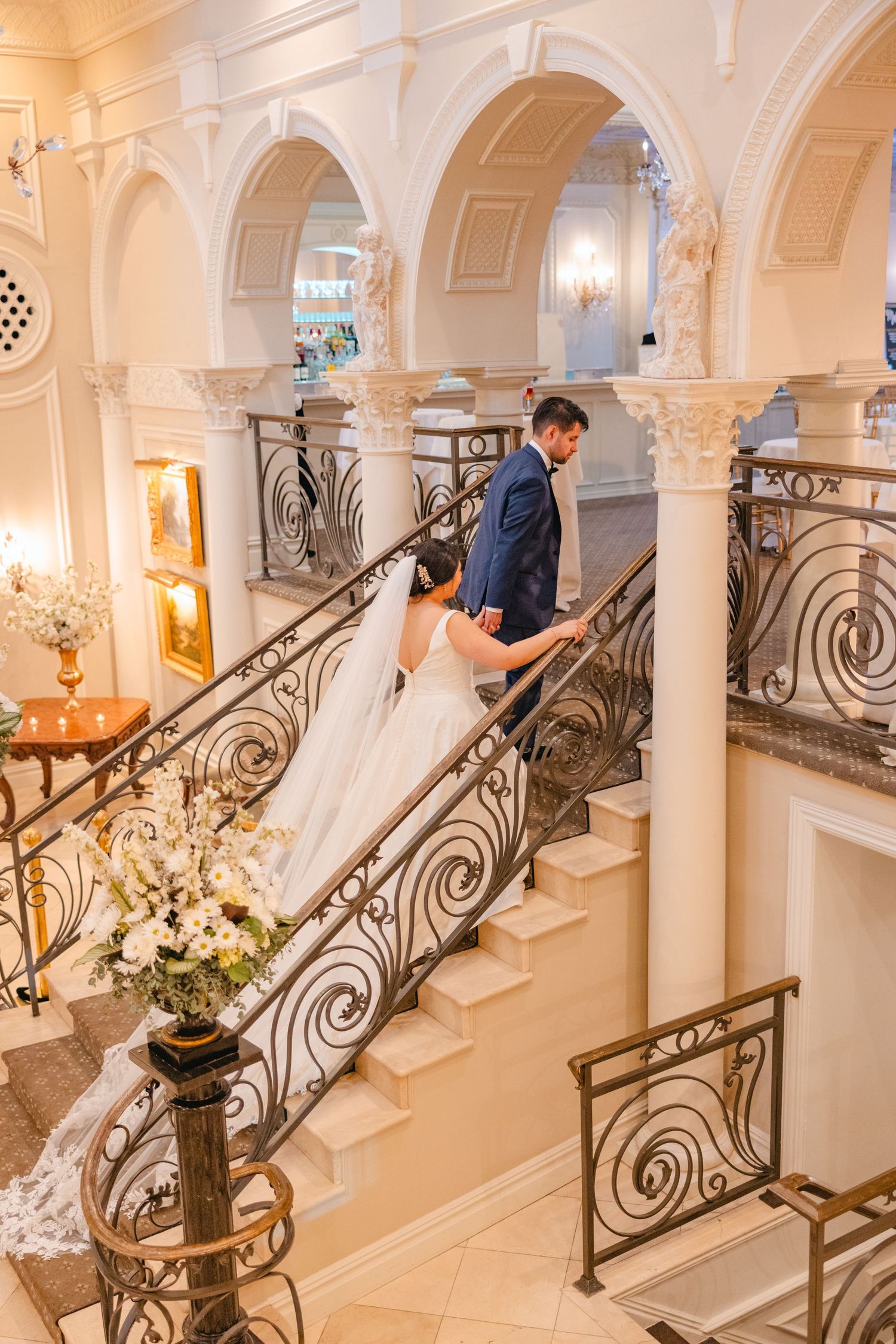 Bride and groom ascending a grand staircase with ornate iron railings in a luxurious wedding venue.