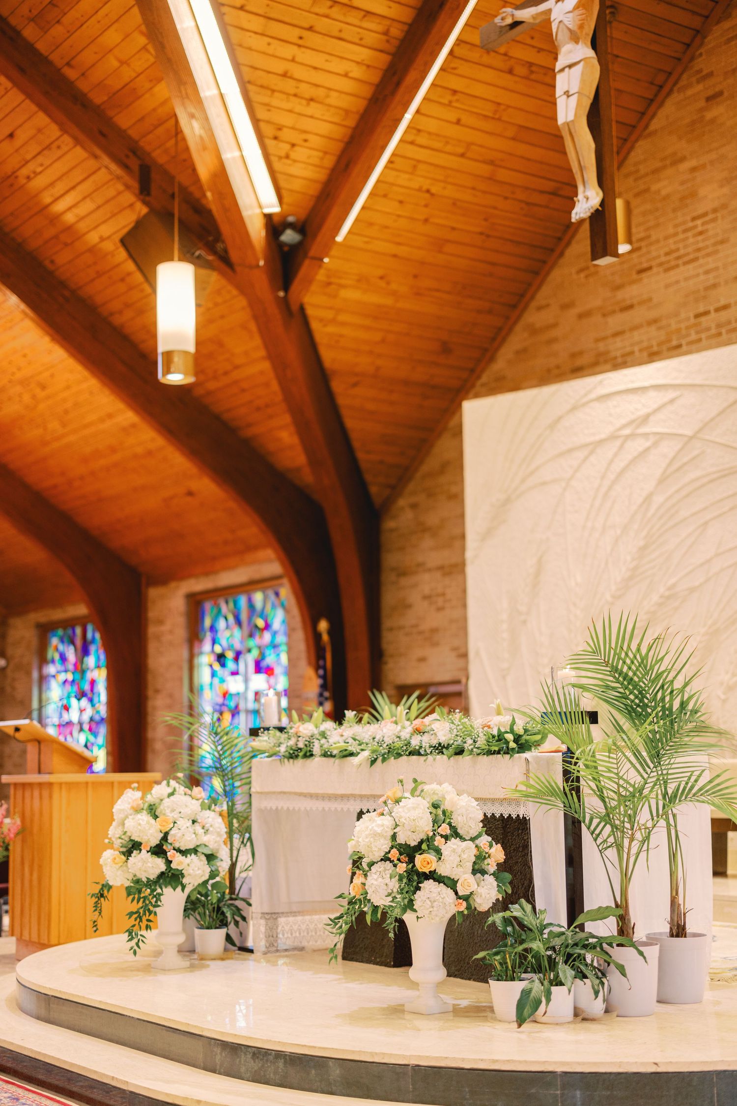 Church altar decorated with elegant white floral arrangements, tropical plants, and a crucifix above the altar.