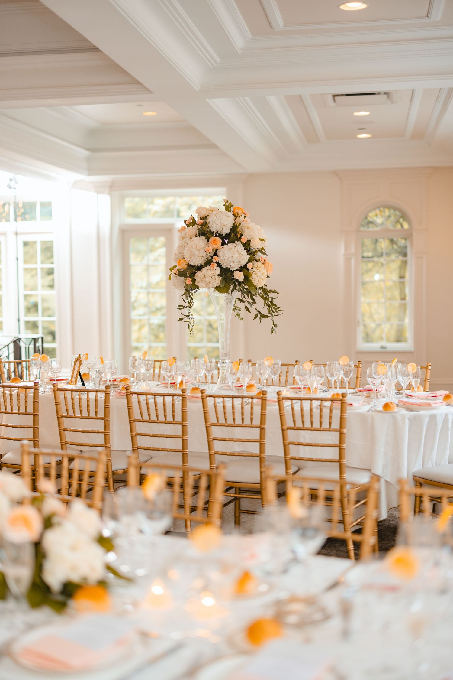 Elegant wedding reception hall with gold chiavari chairs, white floral centerpieces, and peach accents on white linen tables.