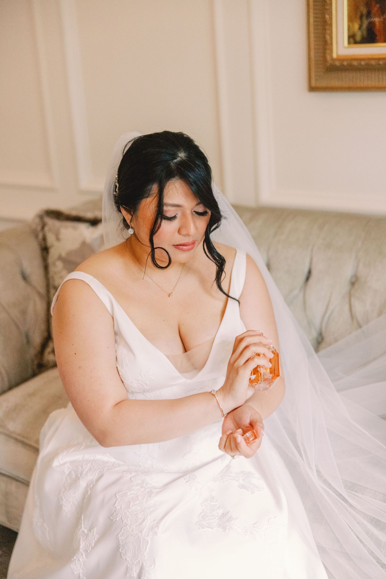 Bride in white wedding gown applying perfume to her wrist while seated on an elegant sofa.