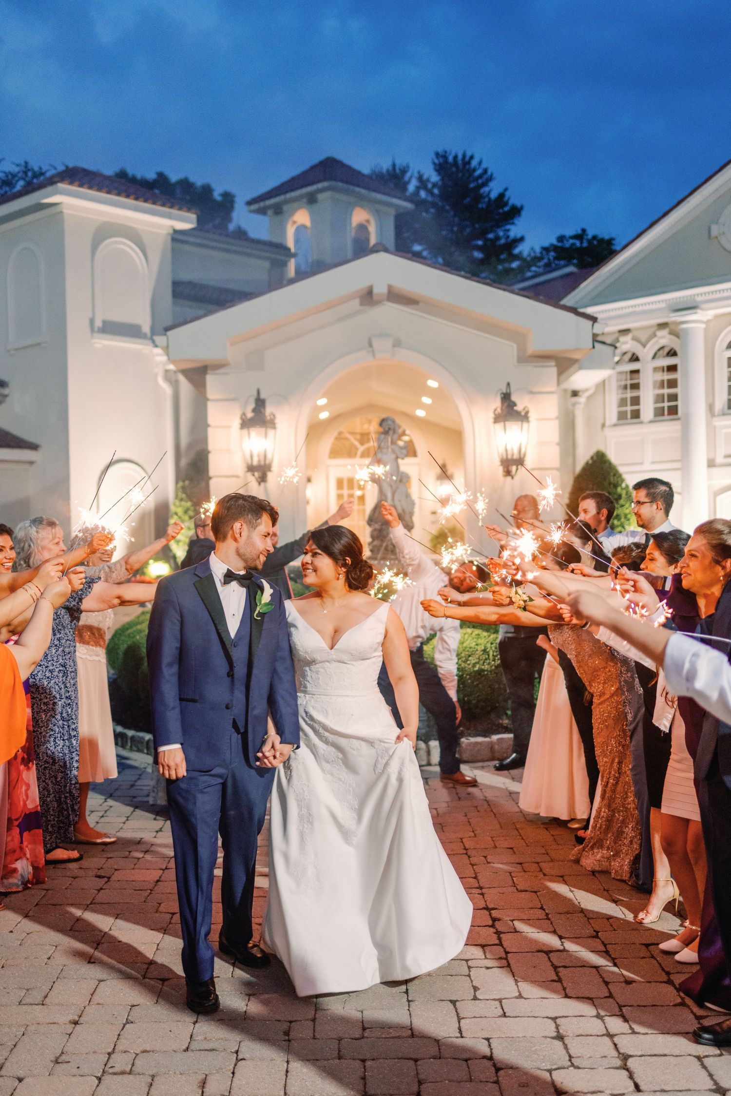 Bride and groom exit through a sparkler tunnel outside a white wedding venue at night.