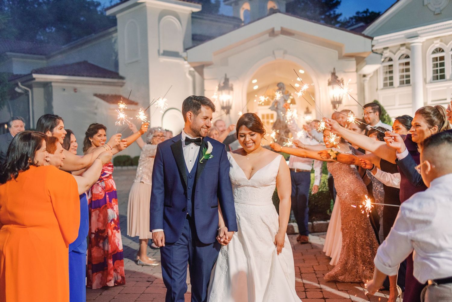 Bride and groom walking through sparkler send-off with guests celebrating outside a grand wedding venue.