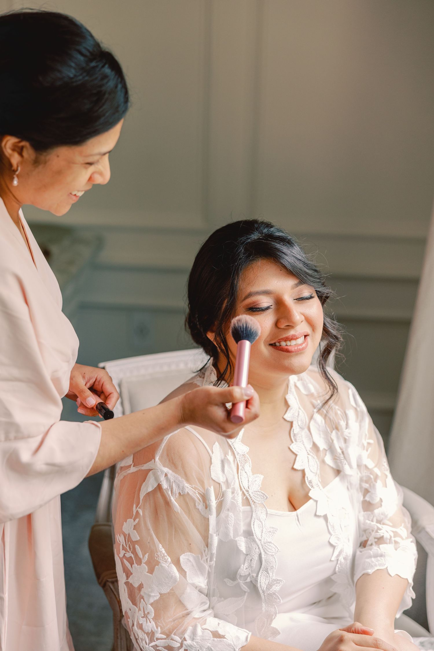Smiling bride in lace robe gets makeup applied with brush by bridesmaid in blush robe.