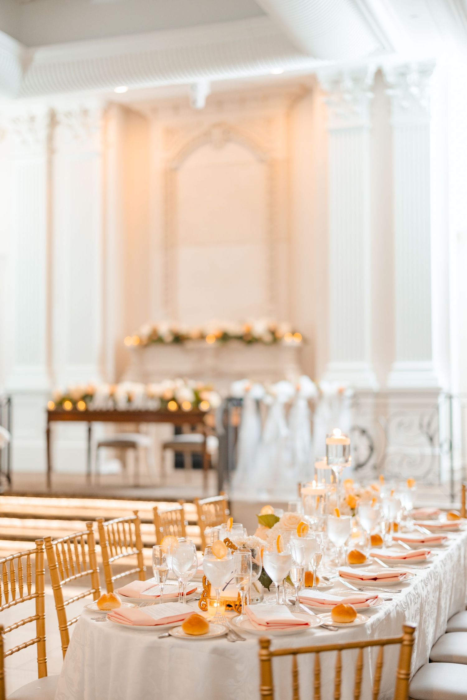 Elegant wedding reception hall with gold chiavari chairs, white tablecloths, pink napkins, and candlelit centerpieces.
