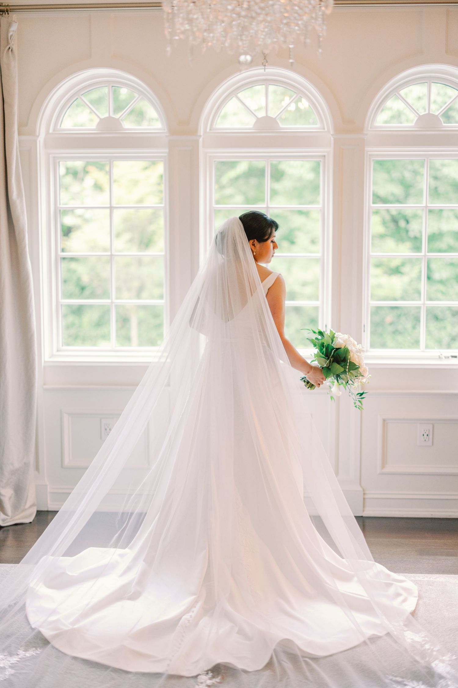 Bride in a flowing white gown and cathedral veil holding a bouquet before arched windows.