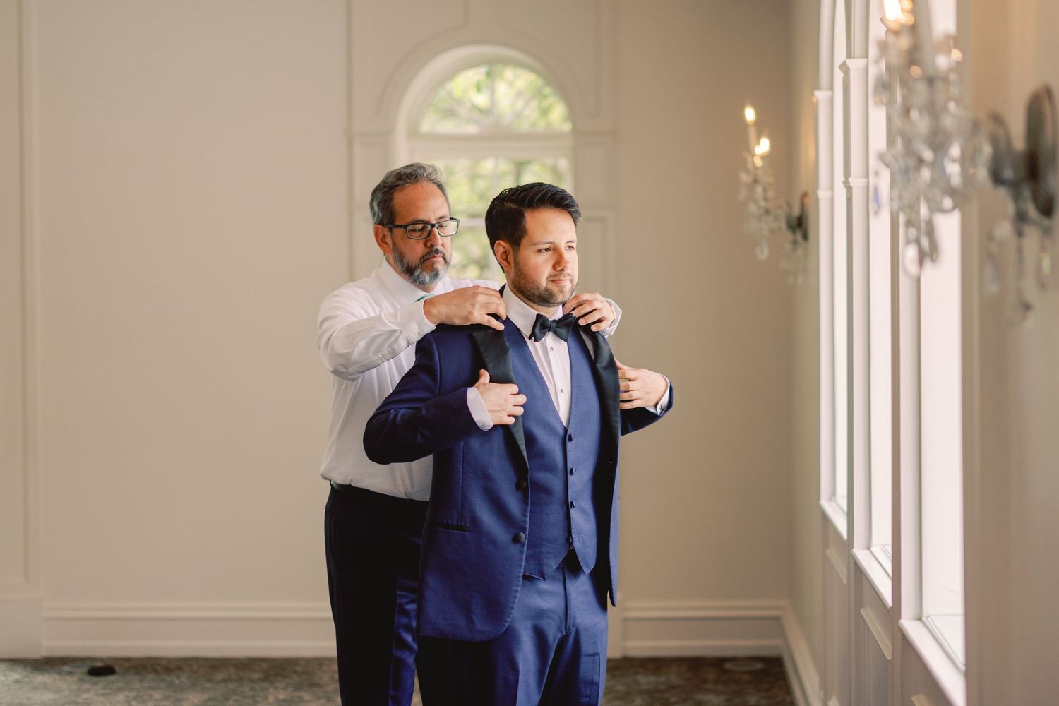 Father helps groom in blue suit adjust bow tie in bright elegant wedding venue room.