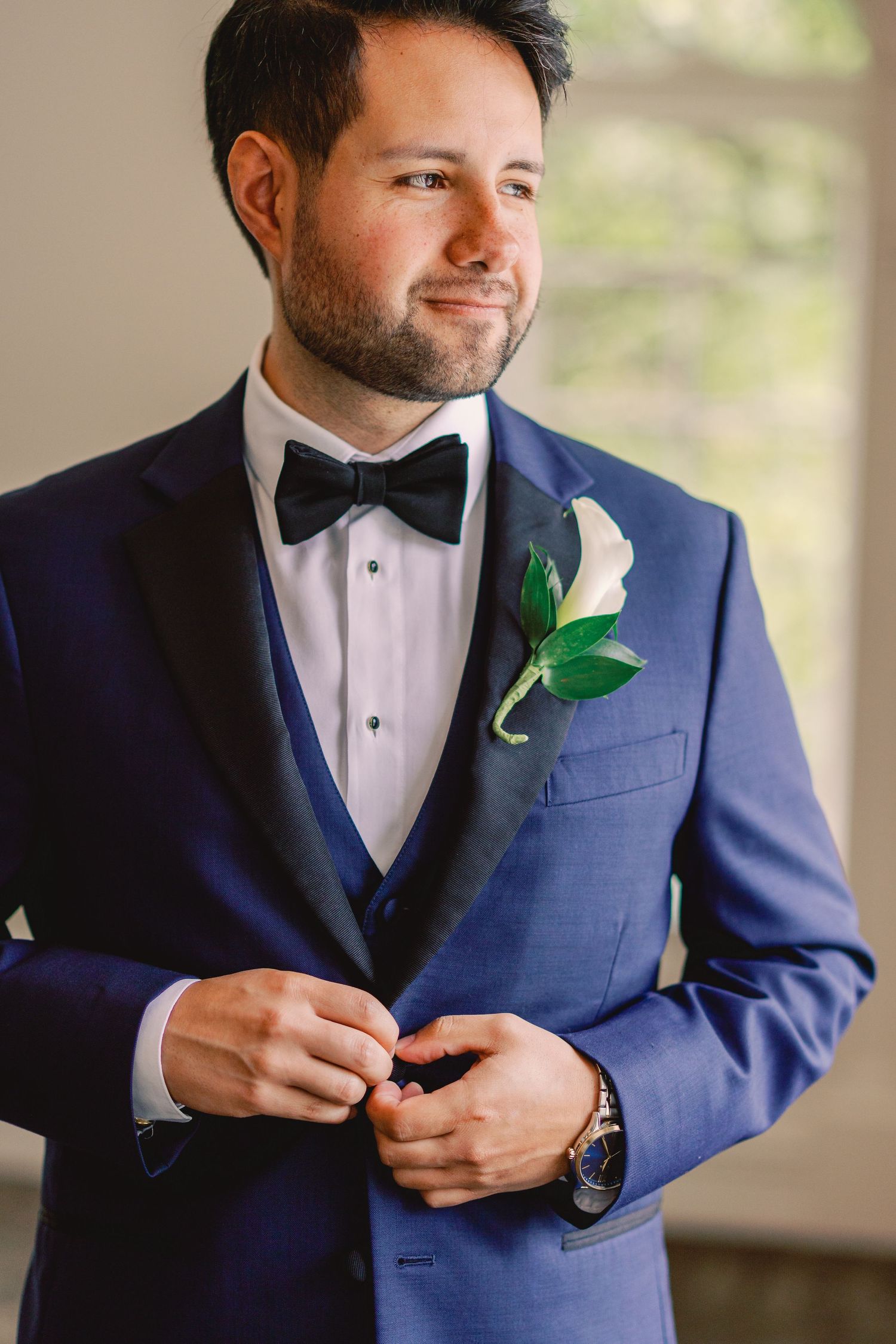Handsome groom in a navy blue tuxedo with black bow tie and white calla lily boutonniere smiles softly.