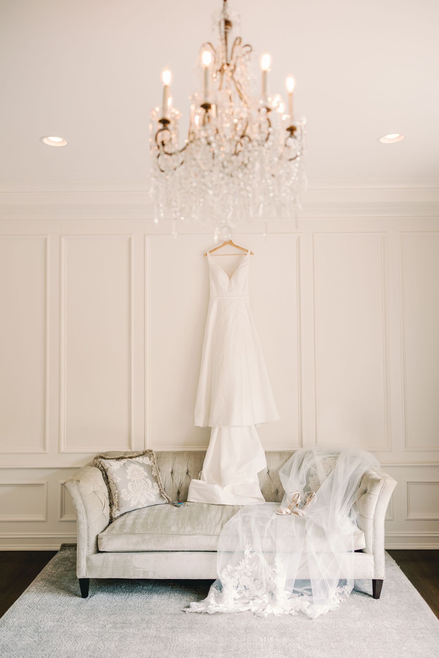 White wedding gown hanging above a tufted sofa with lace veil draped elegantly beneath a chandelier.