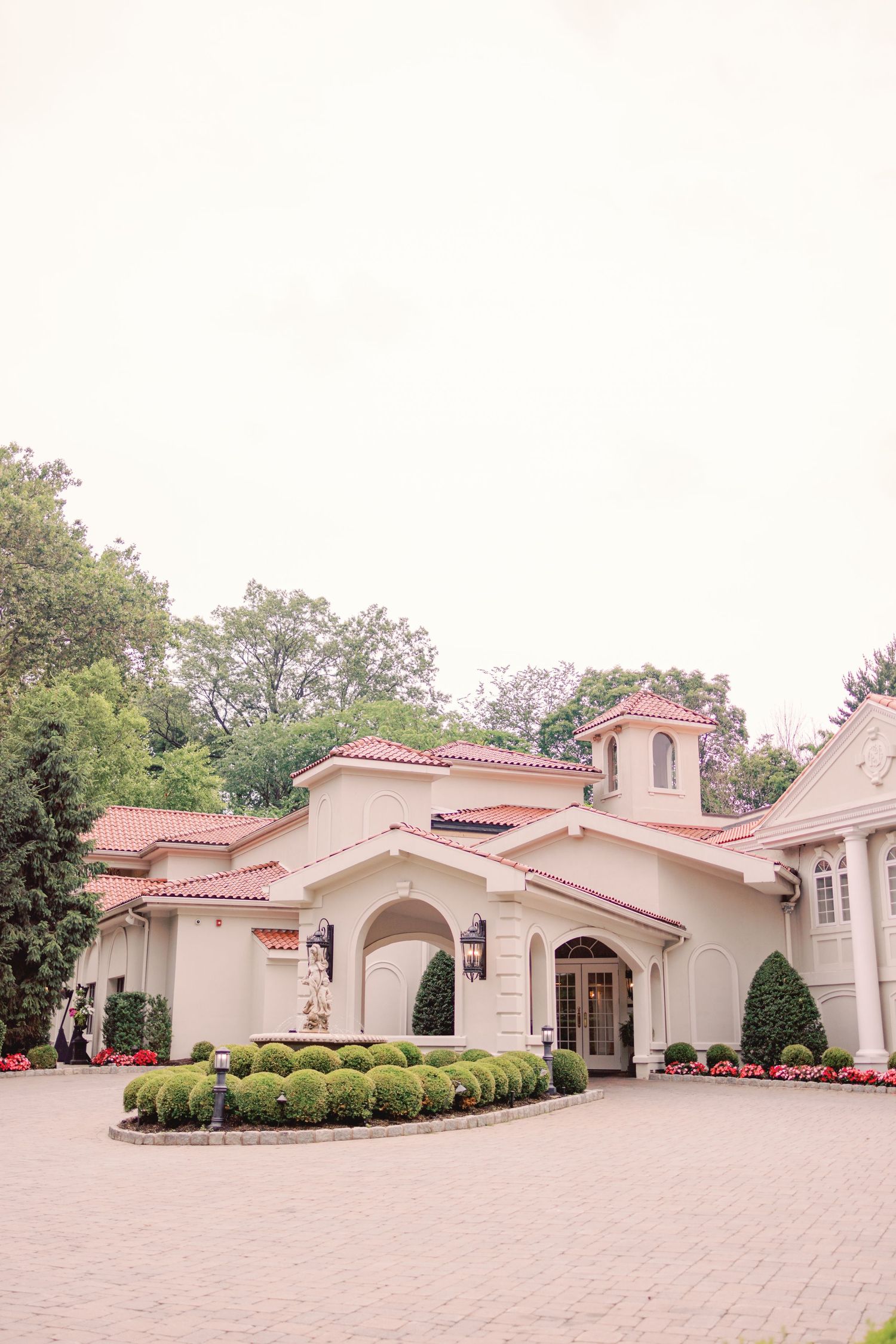 Elegant wedding venue exterior with red-tiled roof, manicured hedges, and blooming flowers.