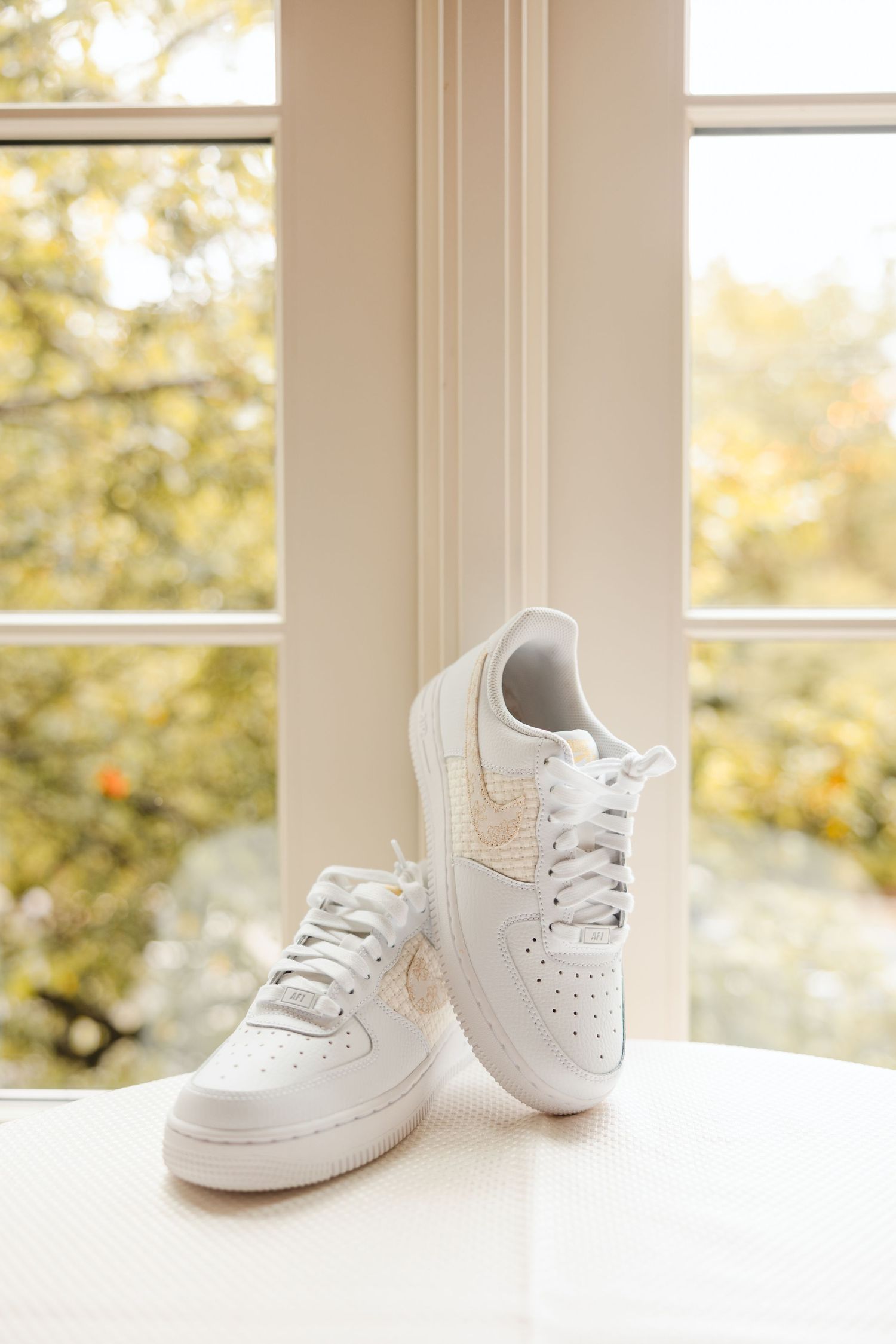 White Nike Air Force 1 custom bridal sneakers displayed on a white surface near a sunlit window.