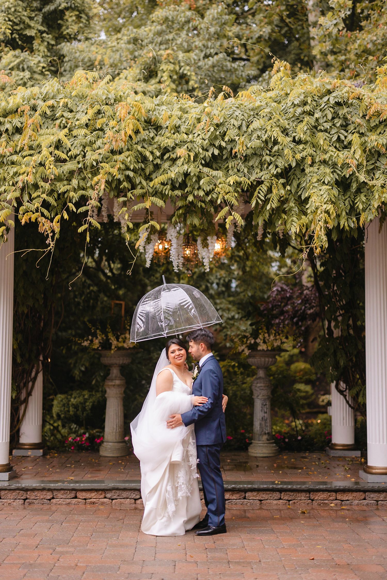 Bride and groom embracing under a clear umbrella beneath a lush green pergola on their wedding day.