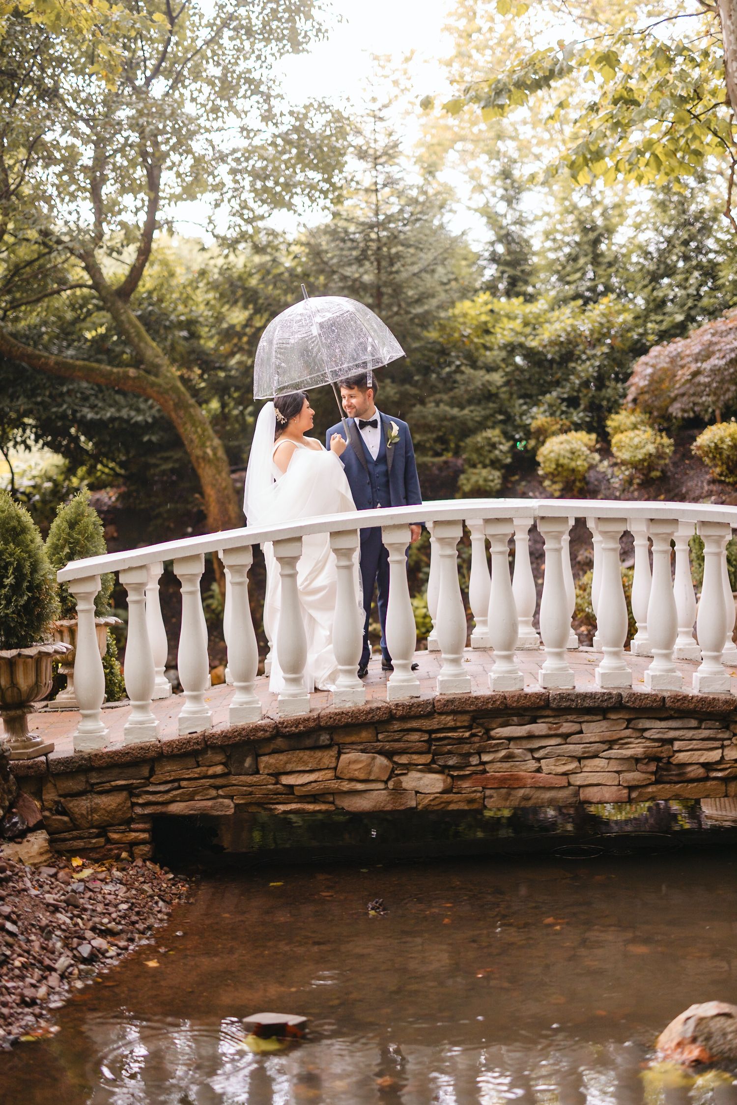 Bride and groom sharing a romantic moment on a stone bridge under a clear umbrella.