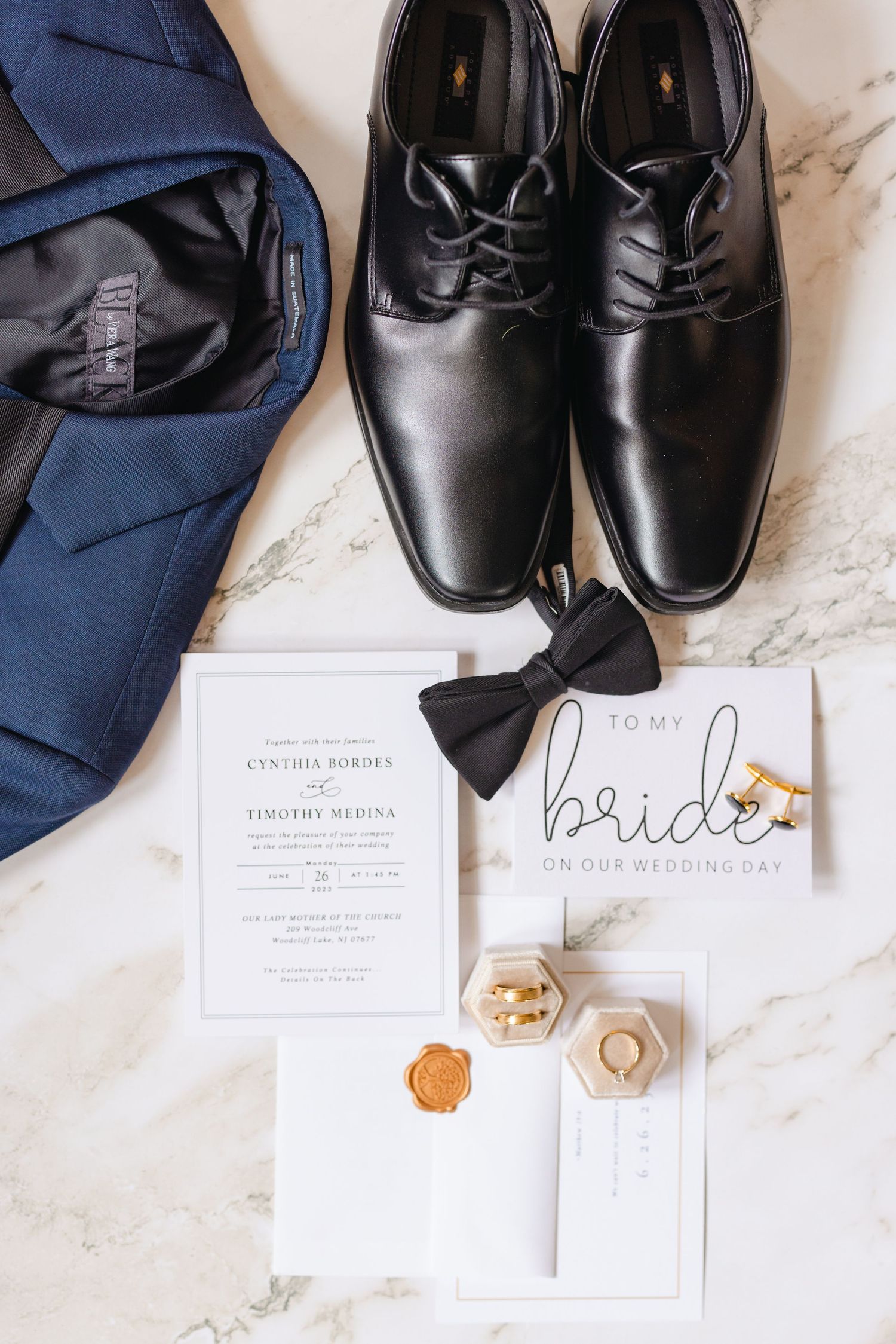 Flat lay of groom wedding details including black oxford shoes, navy suit, bow tie, rings, and invitation.