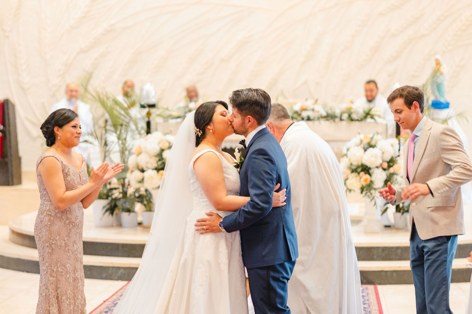 Bride and groom share their first kiss at the altar surrounded by white floral arrangements and wedding party.