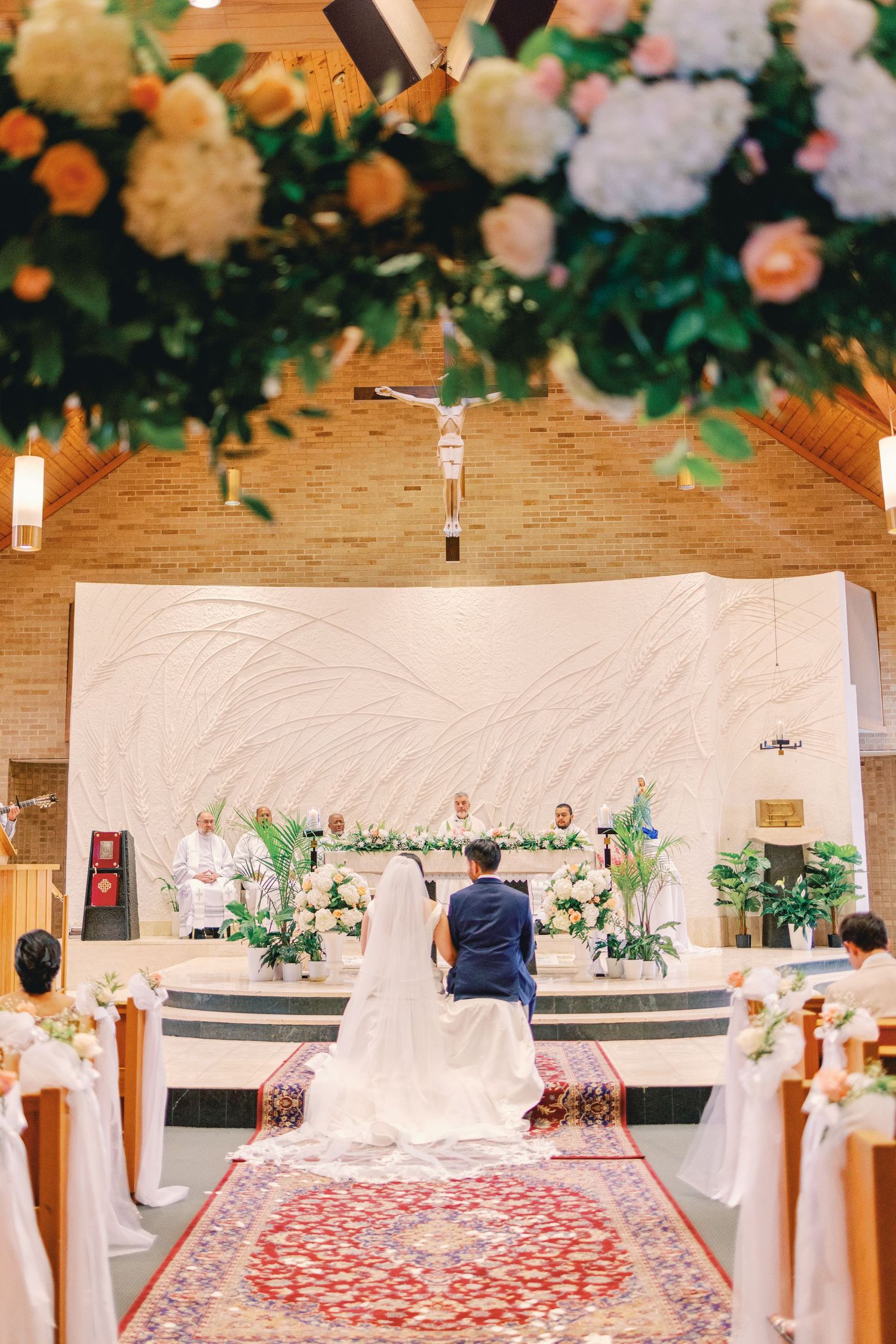 Bride and groom kneeling at the altar during a Catholic wedding ceremony adorned with white floral arrangements.