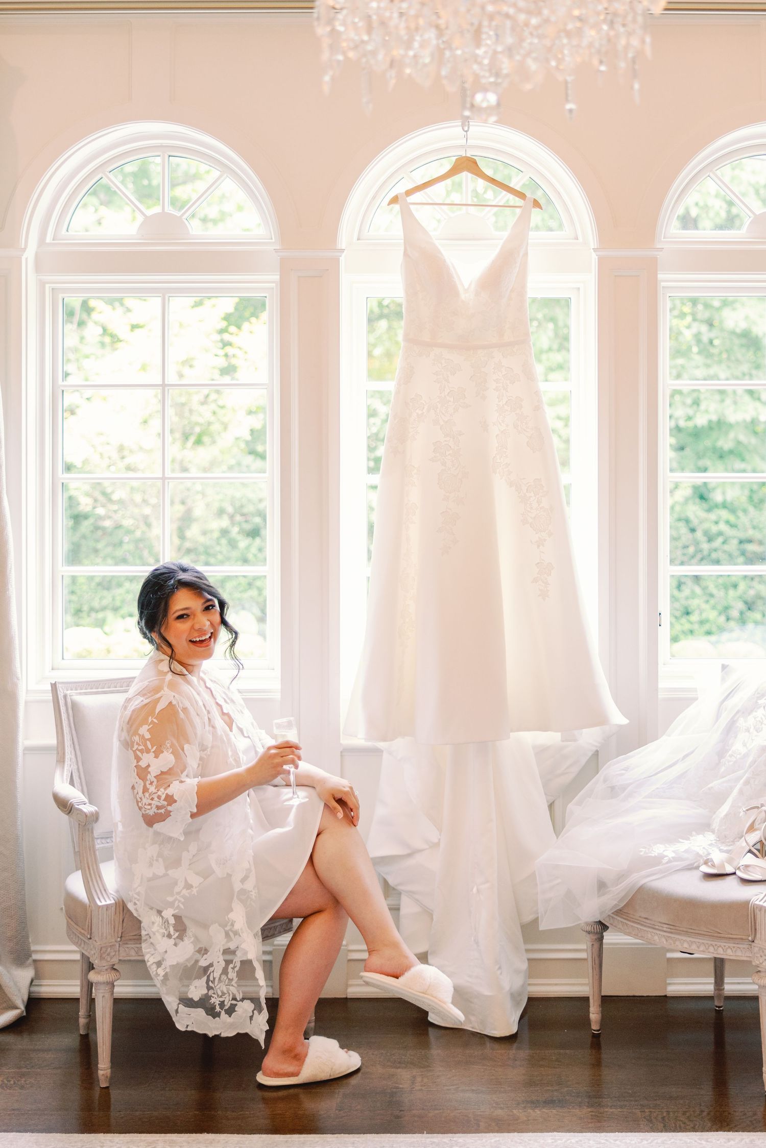 Radiant bride in lace robe sits smiling before hanging wedding dress in bright sunlit room.