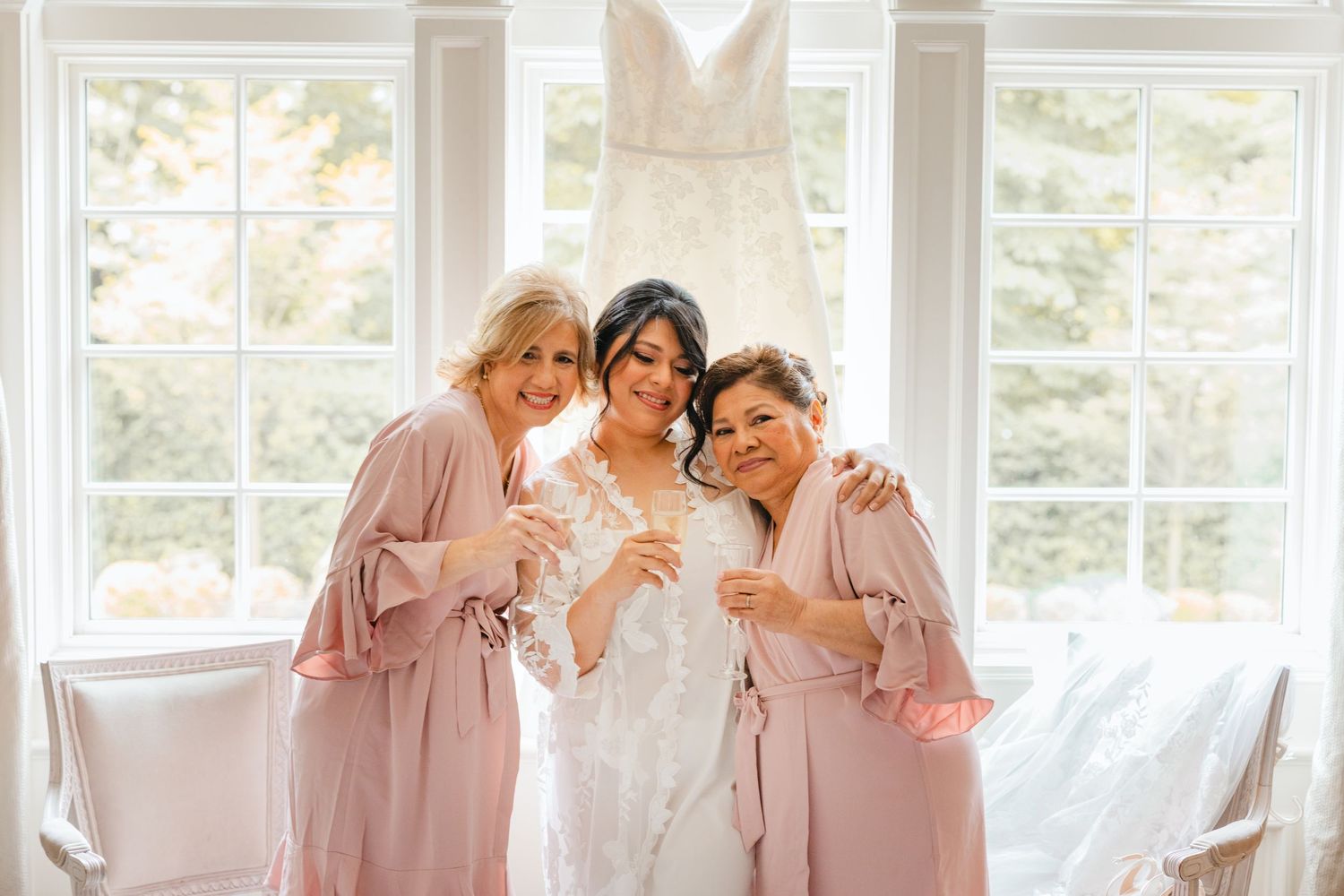 Bride in lace robe poses with two bridesmaids in blush robes holding champagne flutes before wedding.