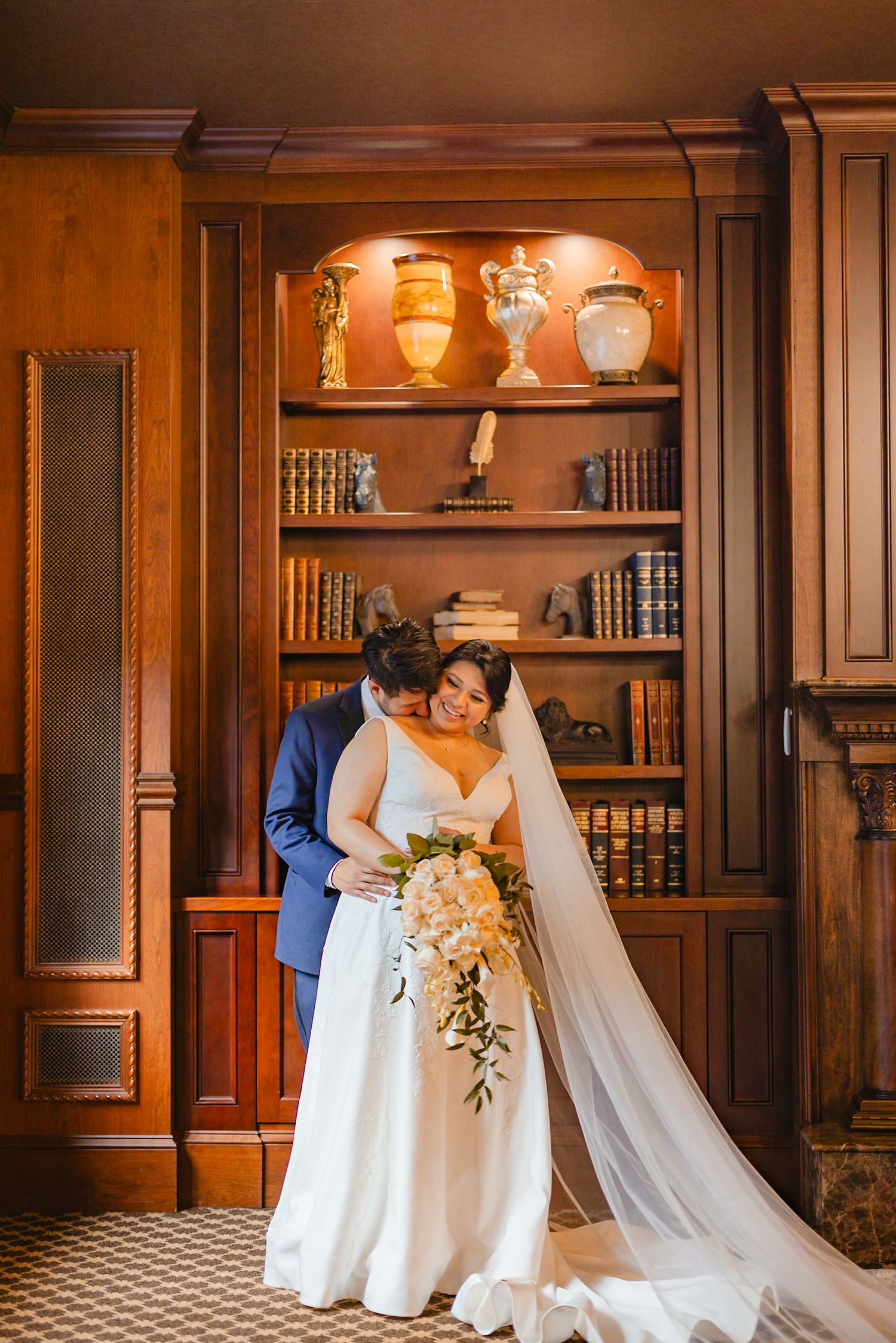 Bride and groom embracing in front of a wooden bookshelf in an elegant library setting.