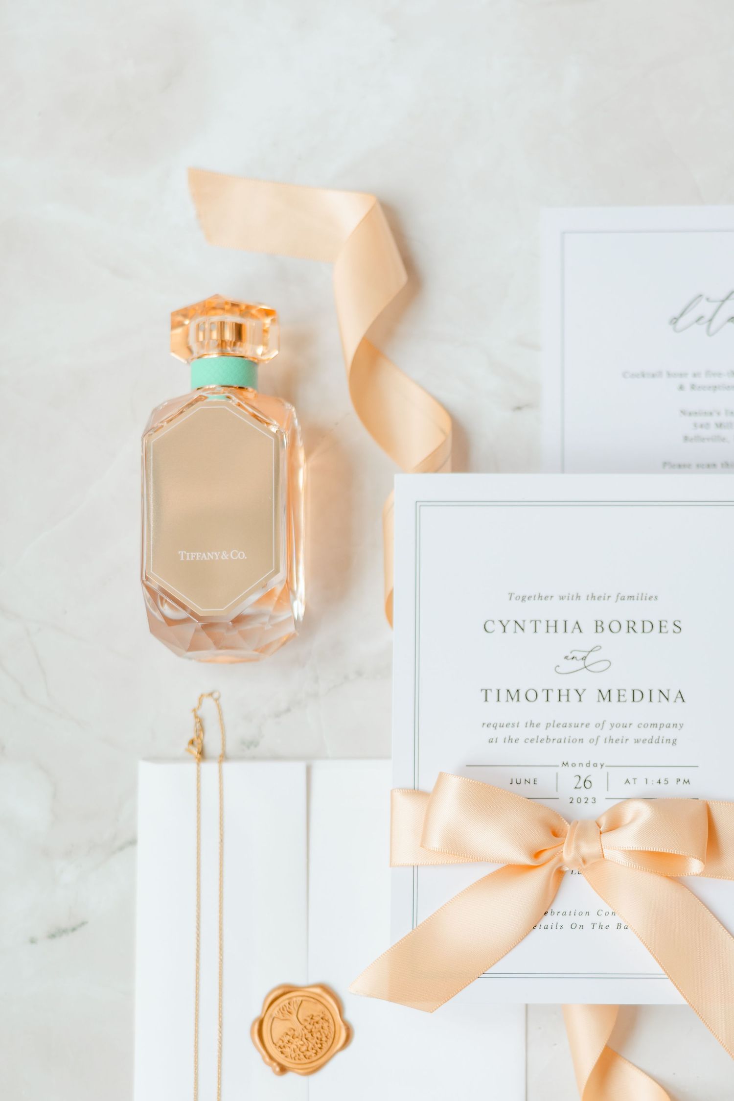 Wedding flat lay featuring a perfume bottle, invitation suite for Cynthia Bordes and Timothy Medina, and peach ribbon.