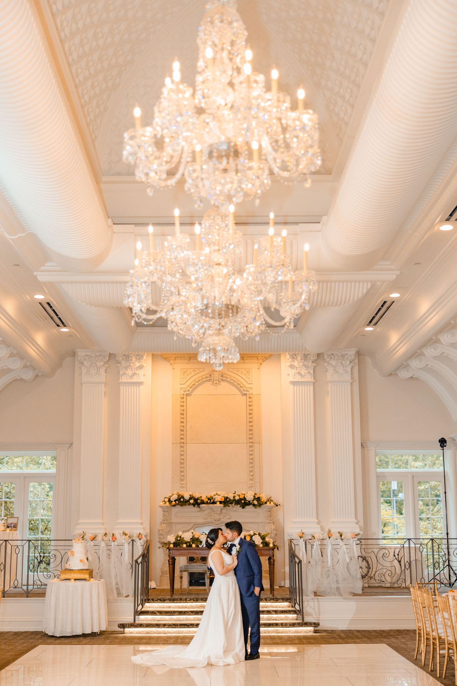Bride and groom share a romantic kiss beneath a grand crystal chandelier in a white ballroom.