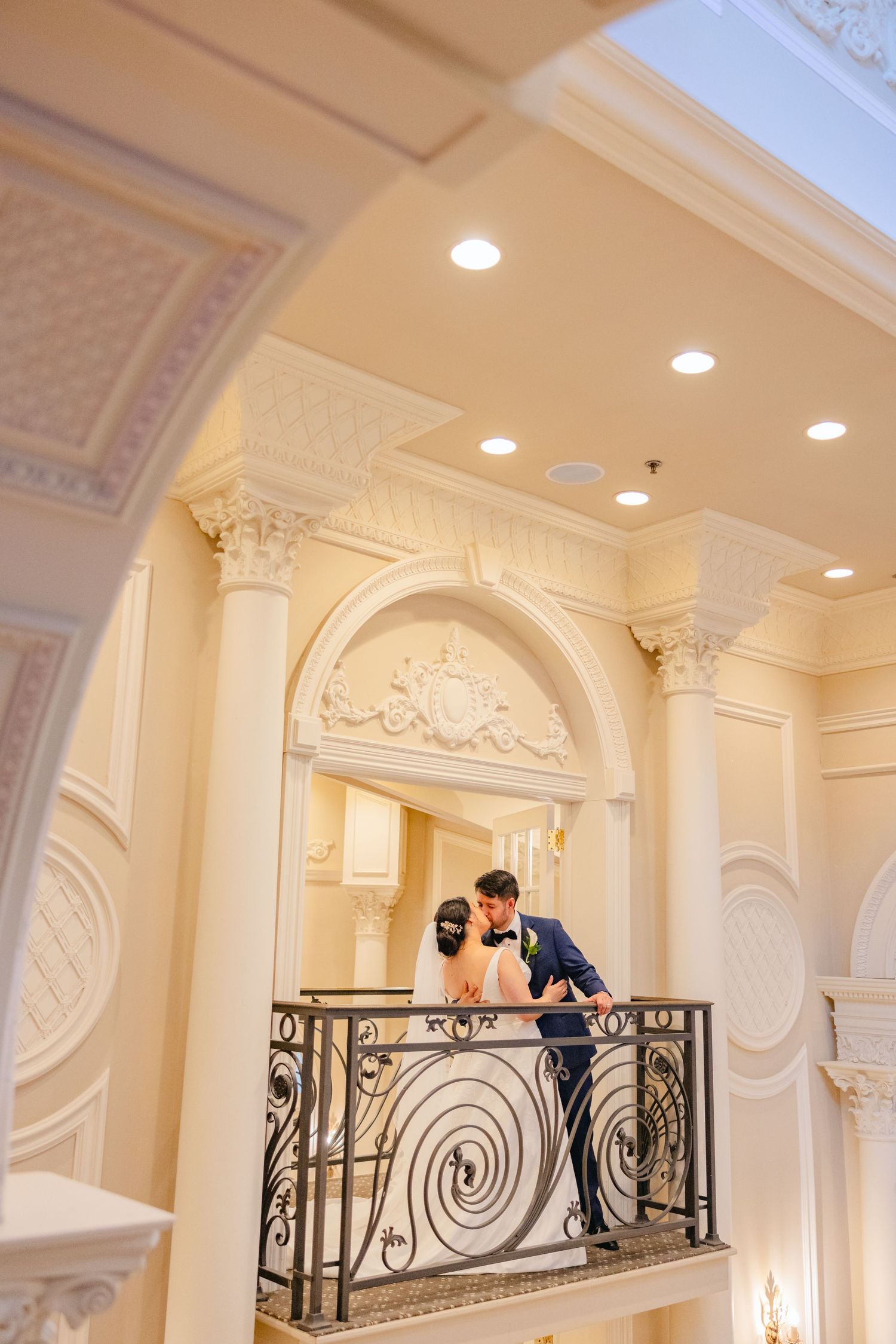 Bride and groom sharing a kiss on an ornate wrought iron balcony inside a grand white wedding venue.