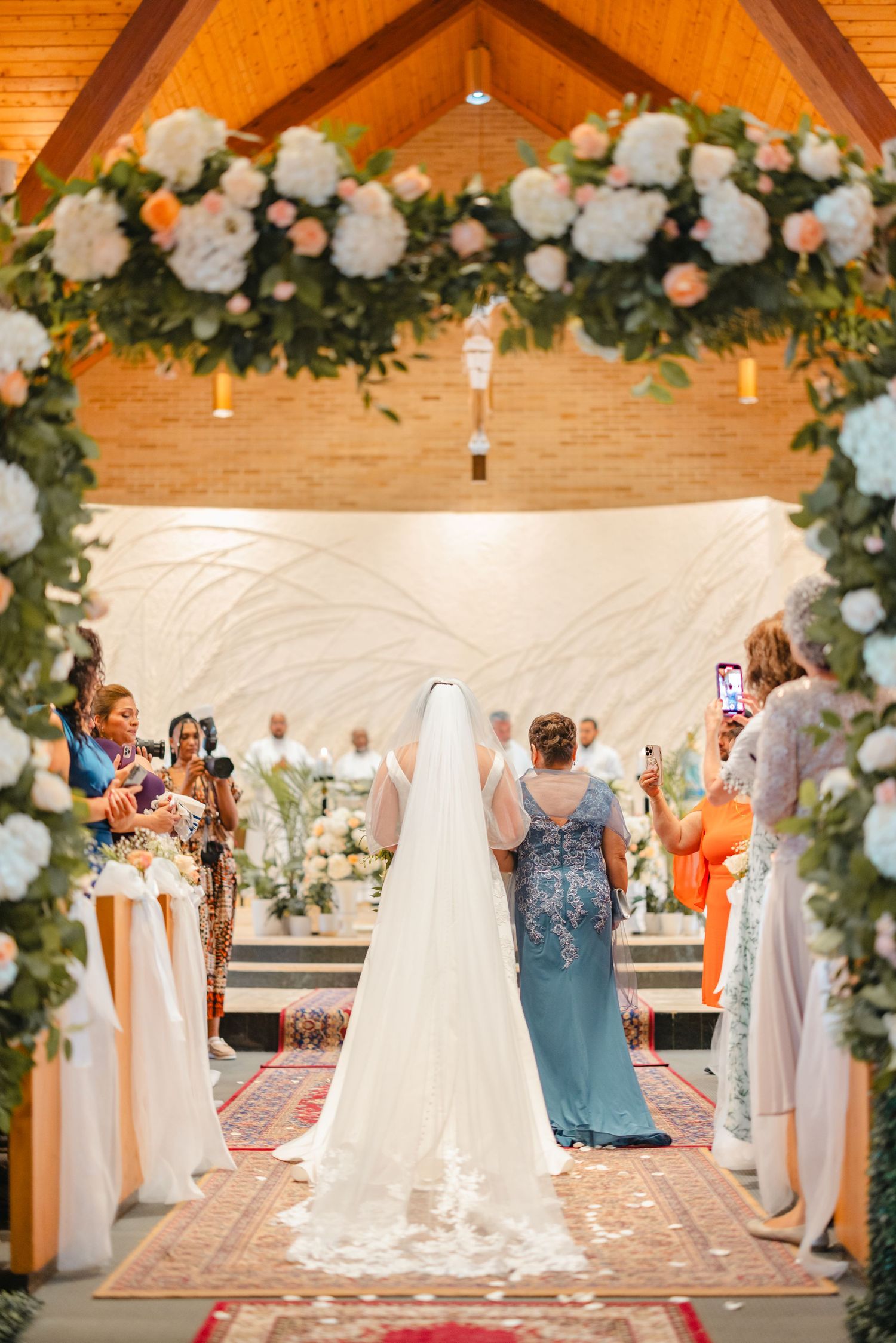 Bride walks down the aisle beneath a stunning floral arch of white and blush roses with her escort.