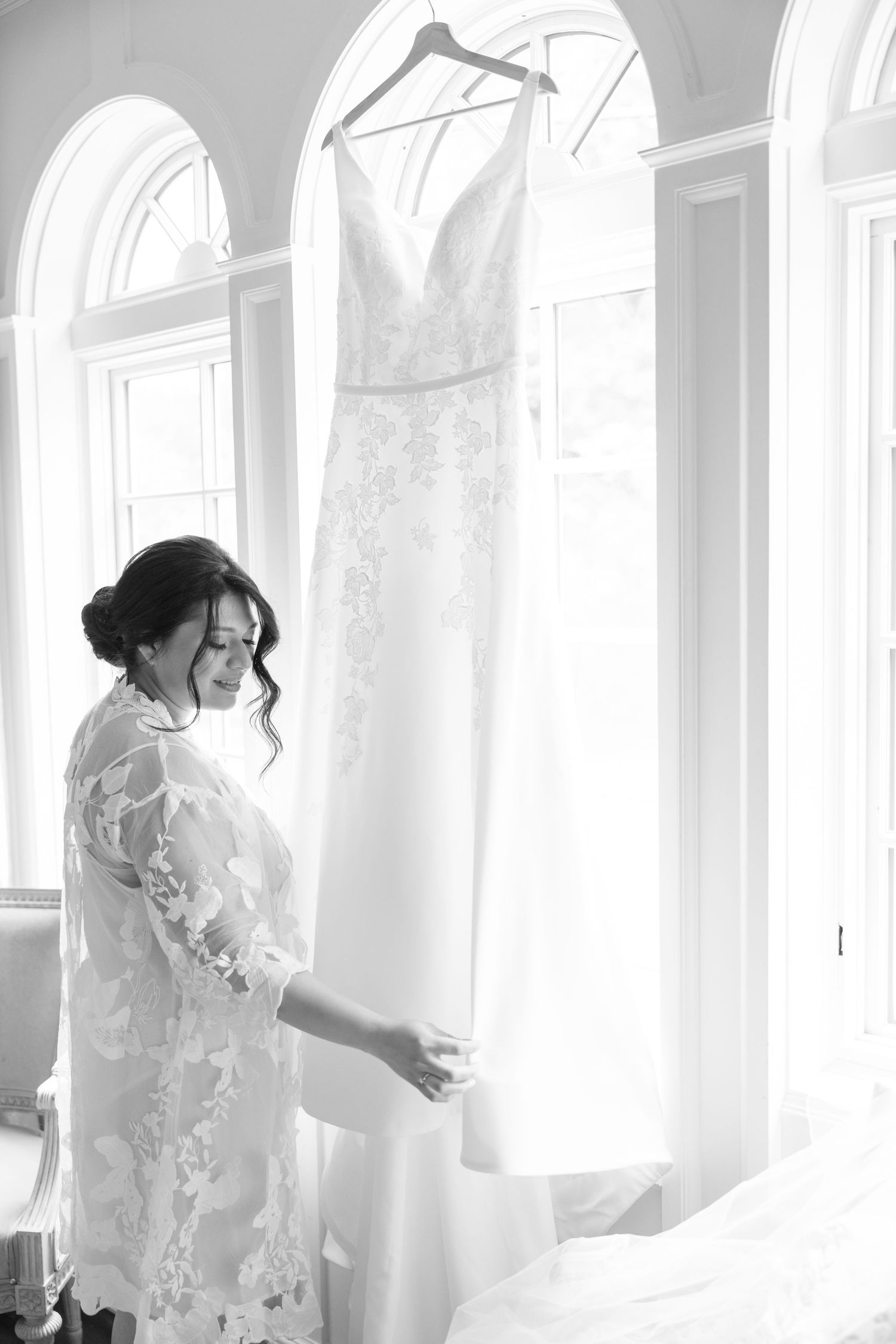 Black and white photo of bride in lace robe admiring her wedding dress hanging by window.