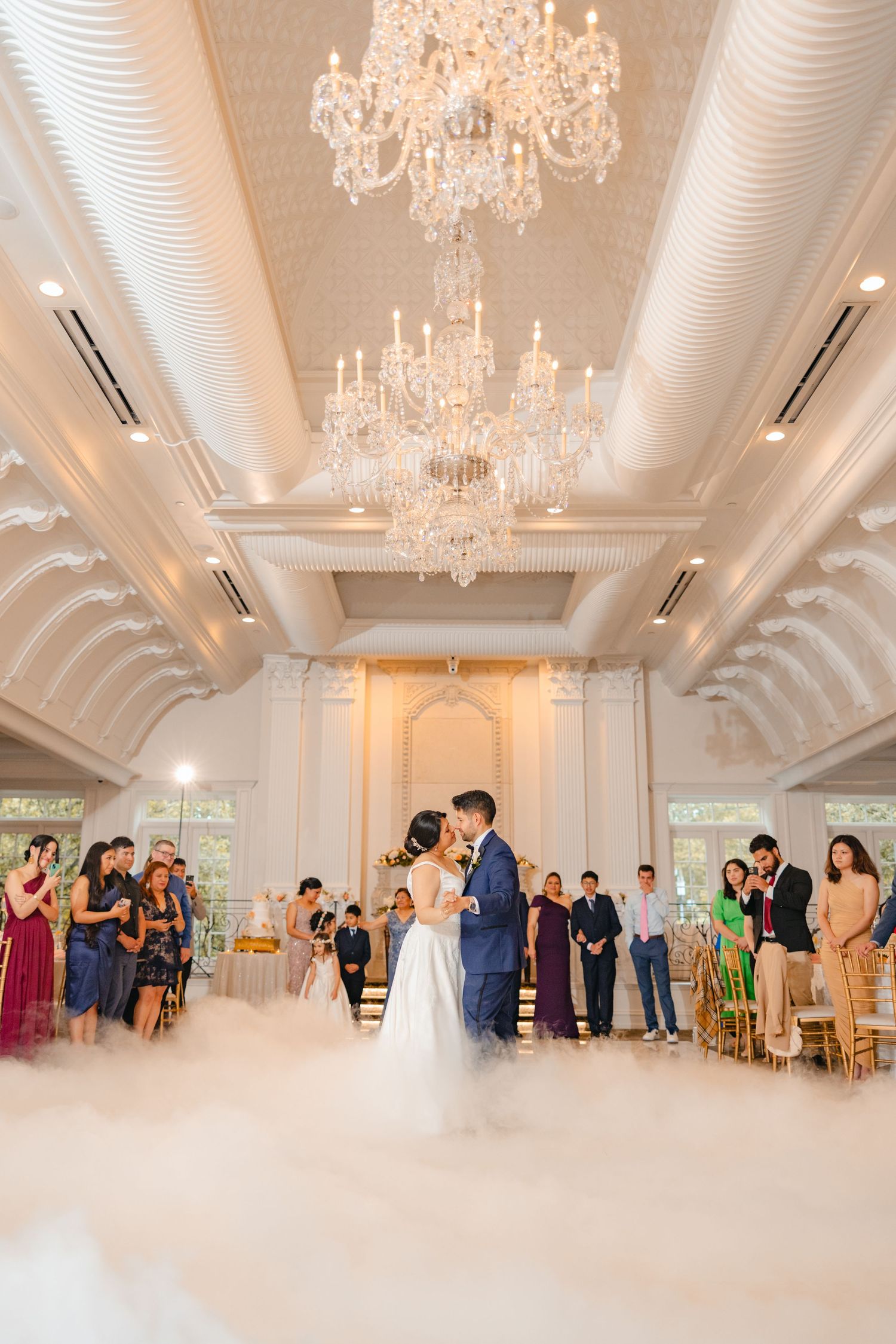 Bride and groom share first dance on a cloud-filled floor beneath crystal chandeliers in an elegant ballroom.