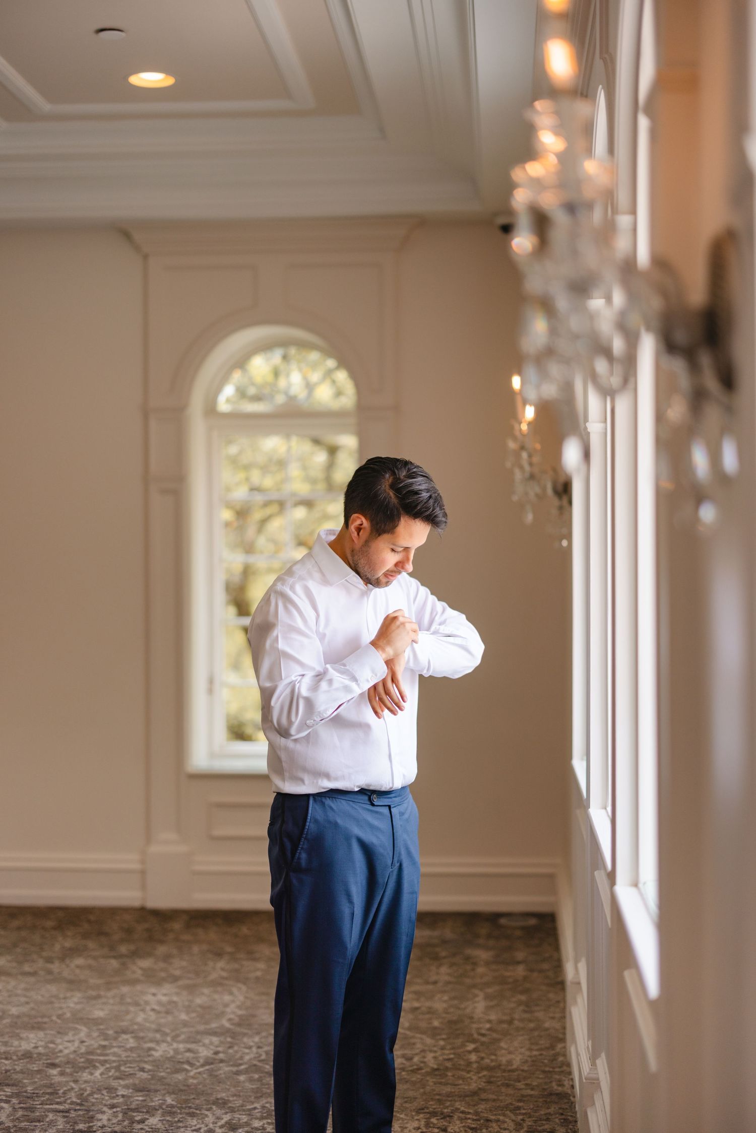 Groom in white dress shirt and navy pants adjusting his cufflinks in an elegant wedding venue hallway.