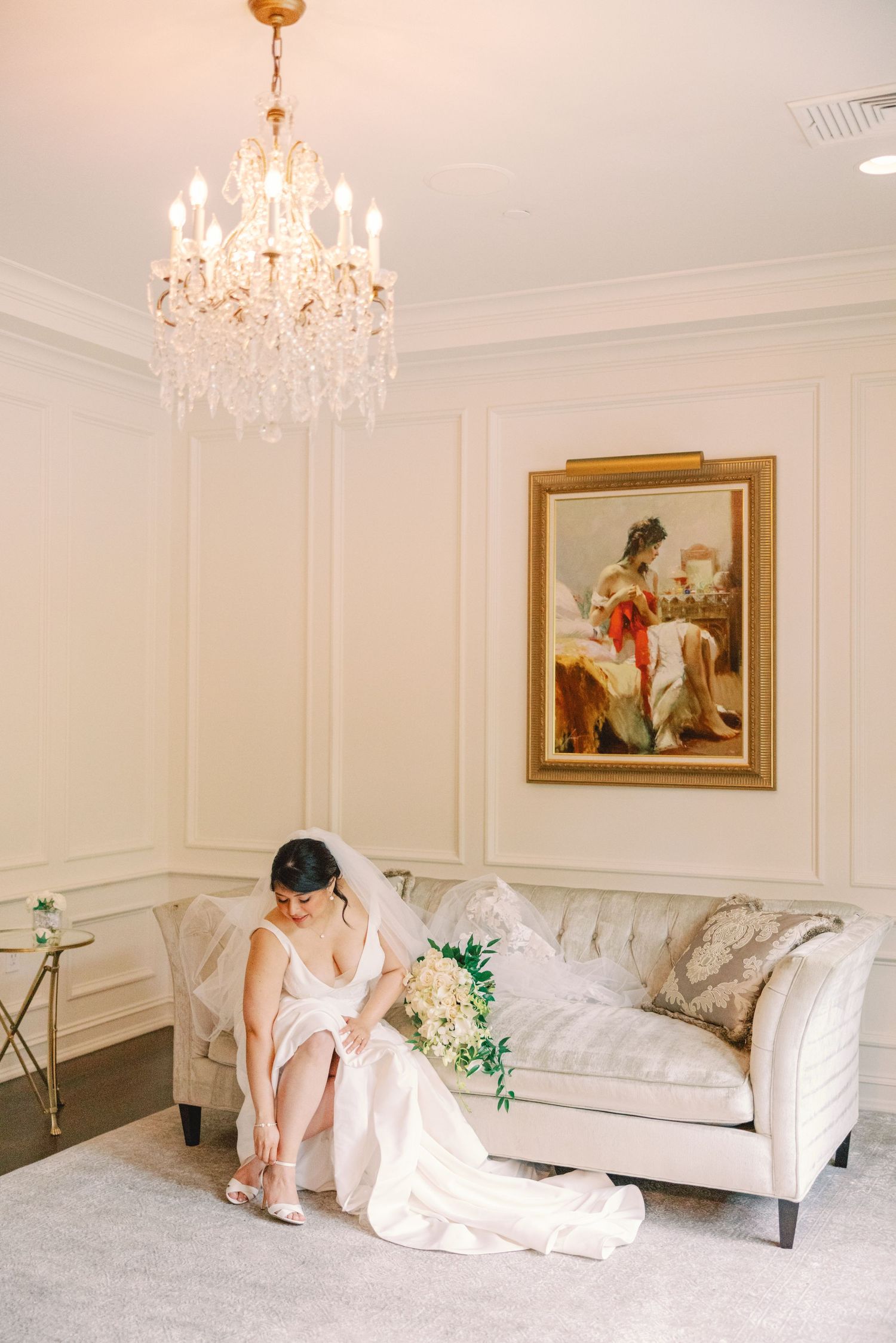 Bride in white gown adjusting her shoes on a tufted sofa beneath a crystal chandelier.