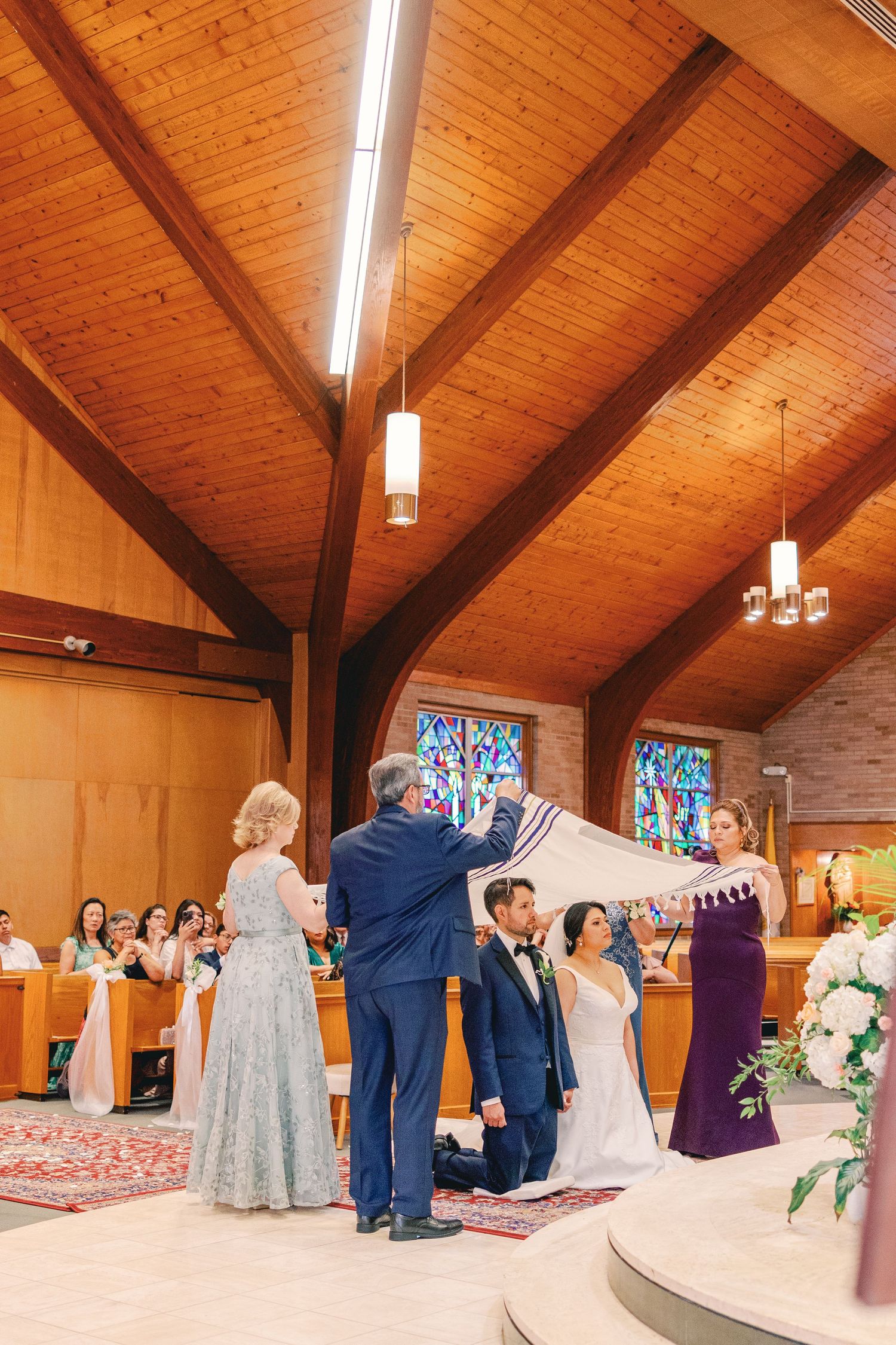 Wedding ceremony with couple kneeling as family holds a veil over them in a wooden church.