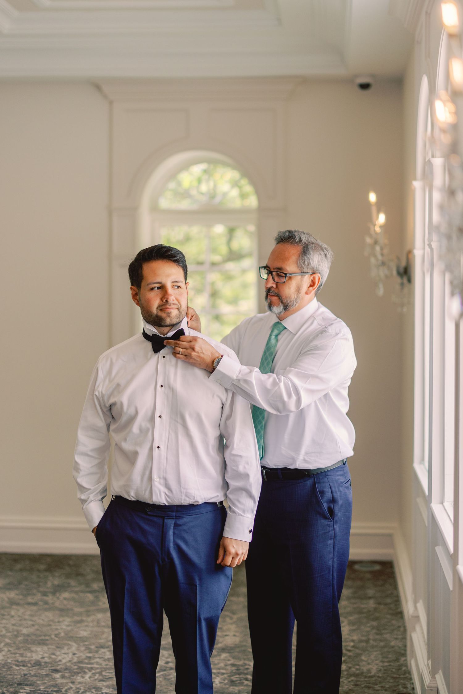 Father helping groom adjust bow tie while both wear white shirts and navy pants in a bright venue.