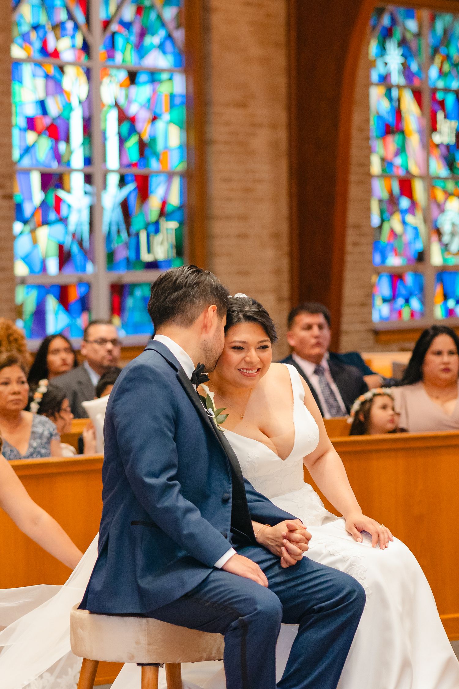 Bride and groom sit together smiling during church ceremony with colorful stained glass windows behind them.