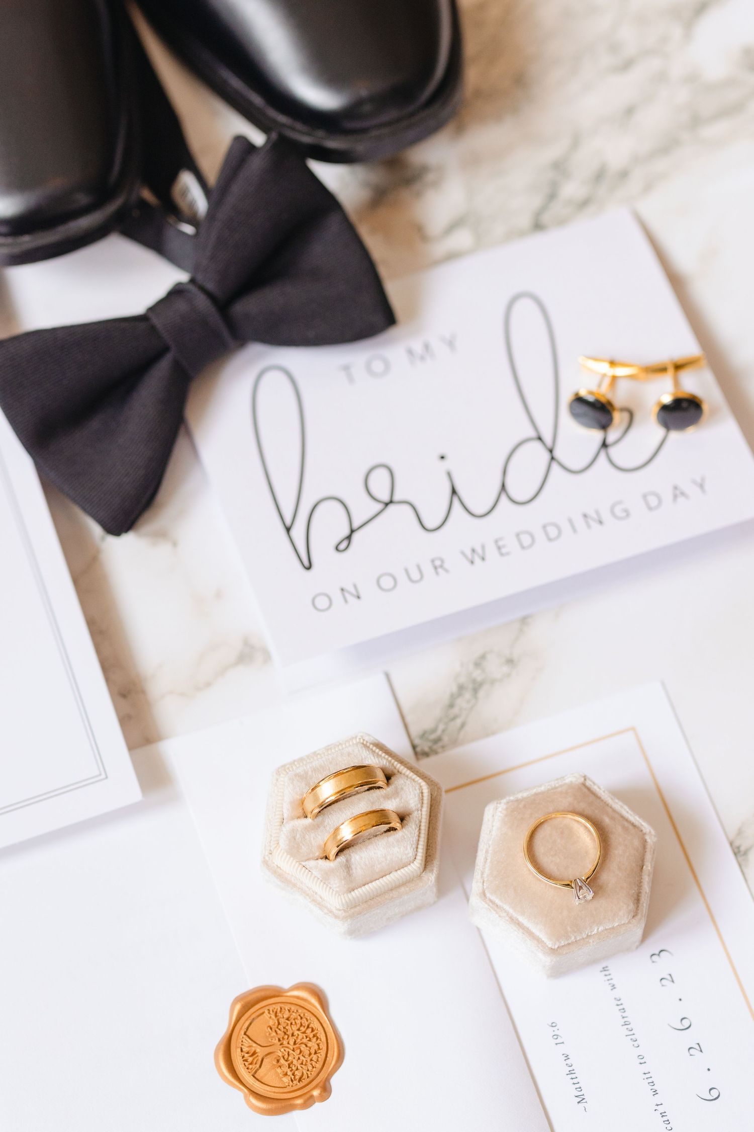 Wedding flat lay featuring gold bands in velvet boxes, bow tie, cufflinks, and bride card on marble.