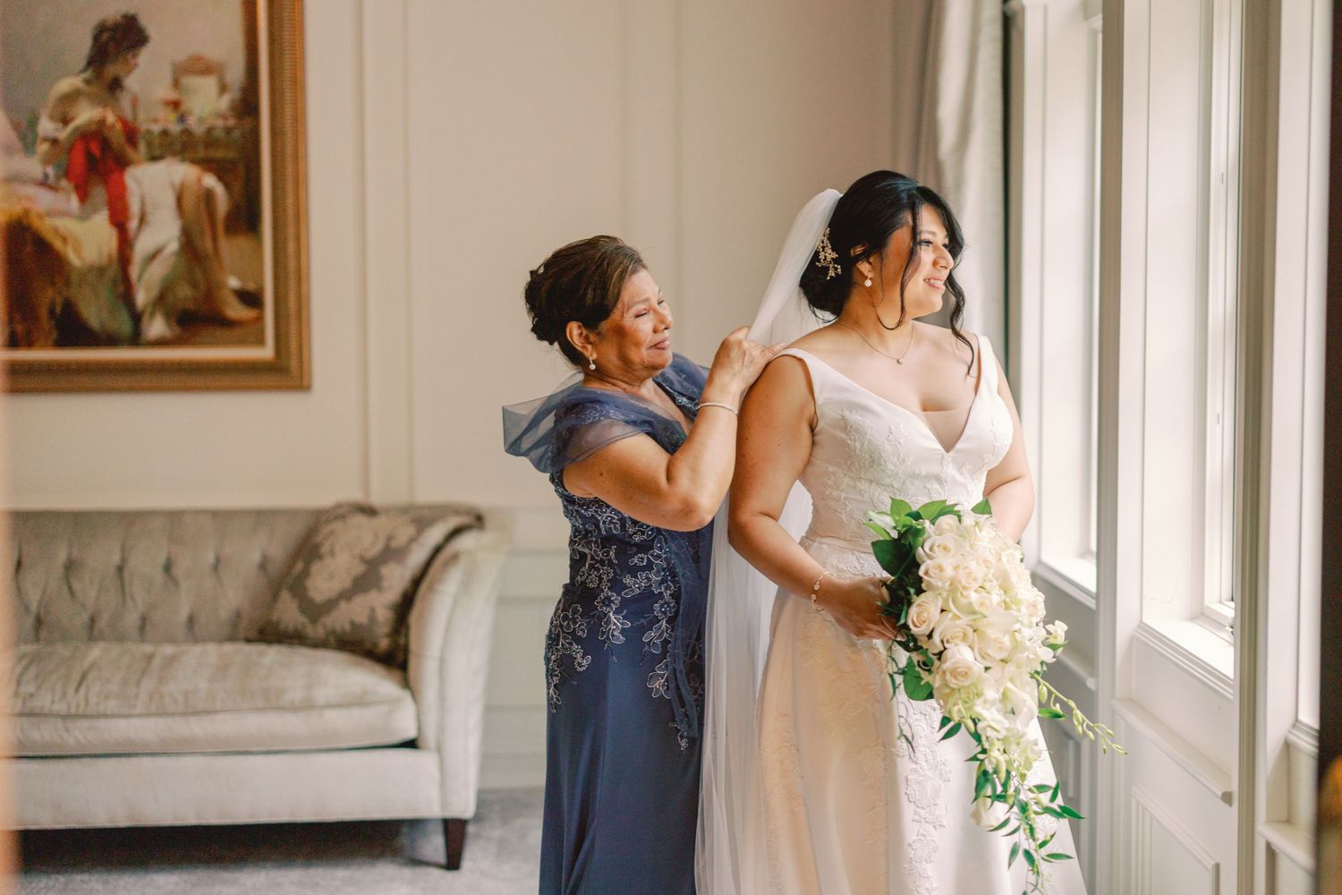 Mother helps bride with her veil near a bright window as bride holds a white bouquet while getting ready.