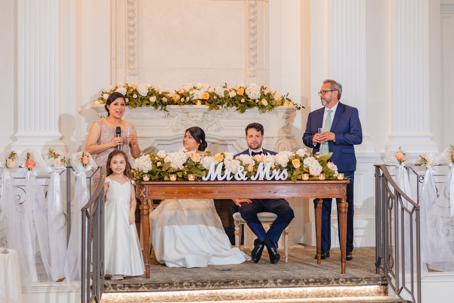 Bride and groom seated at sweetheart table with Mr and Mrs sign during wedding reception toasts.